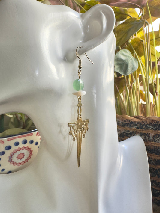 Gold sword earring with green bead on a mannequin ear against a blurred indoor background