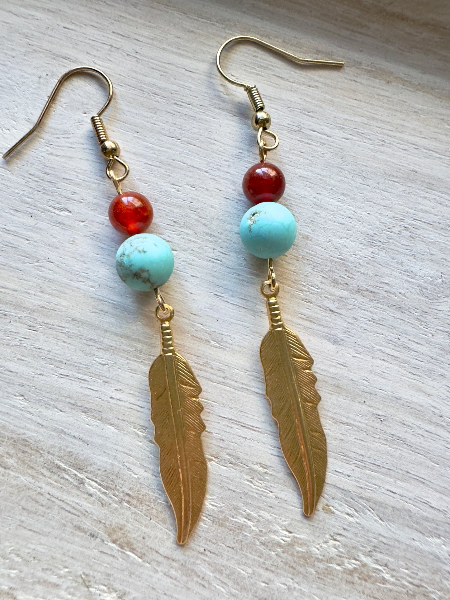 Pair of earrings with gold feather charms and colorful beads on a light wooden surface