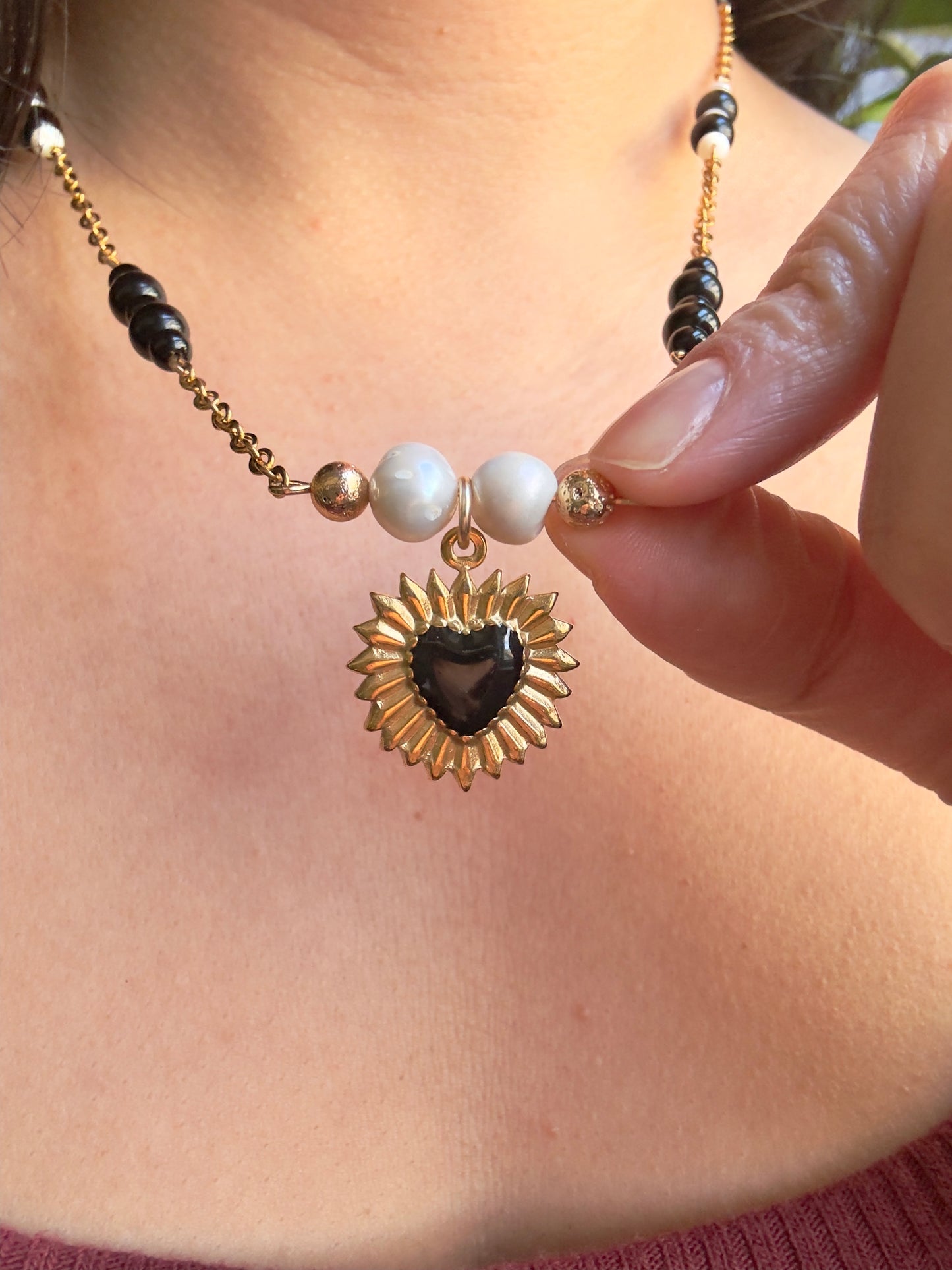 Black heart gold stainless steel pendant on gold stainless steel chain with black jasper and pearl beads, close up of model holding heart pendant