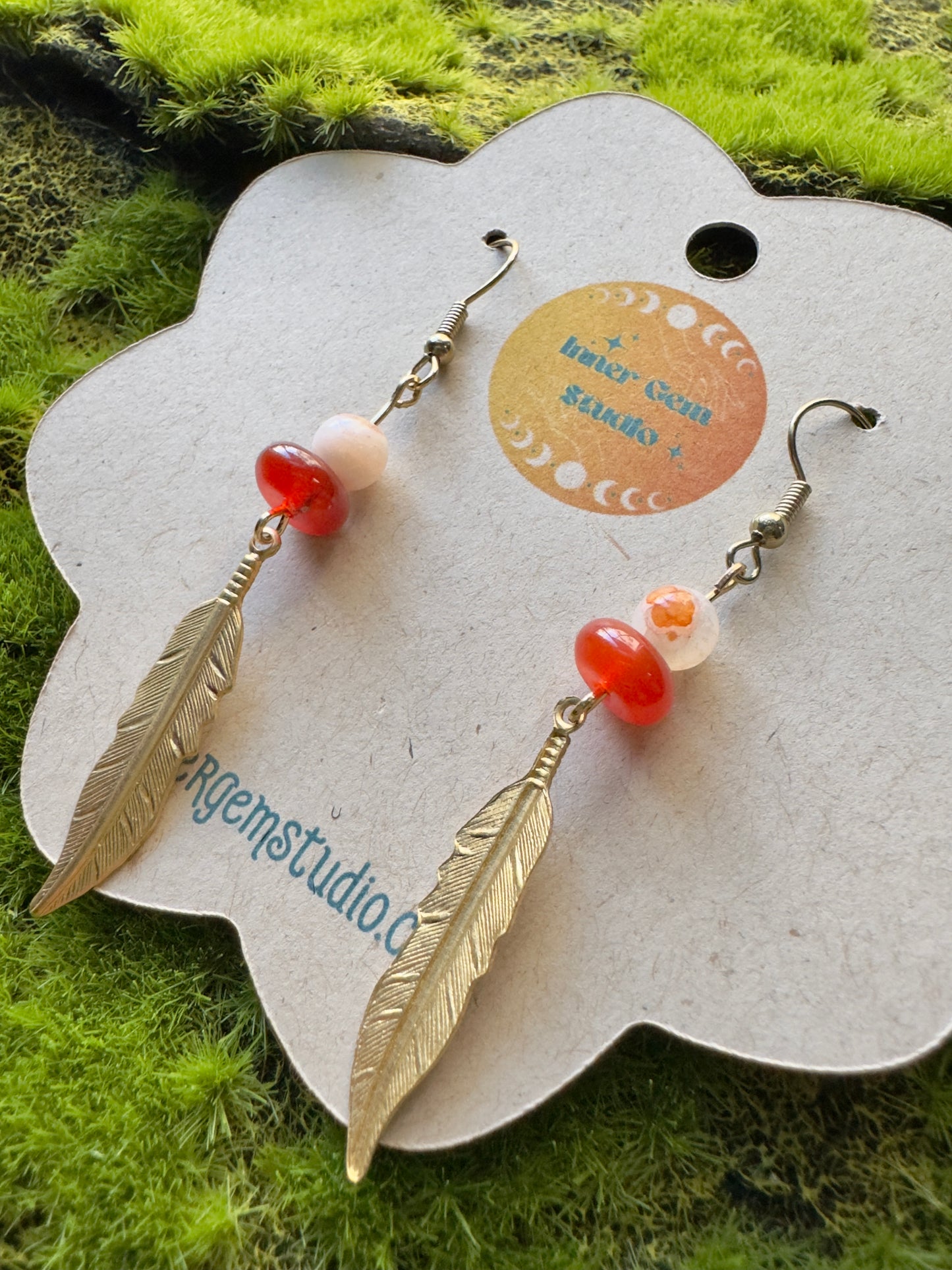 Feather-shaped earrings with red and pink beads on a branded card against a grassy background