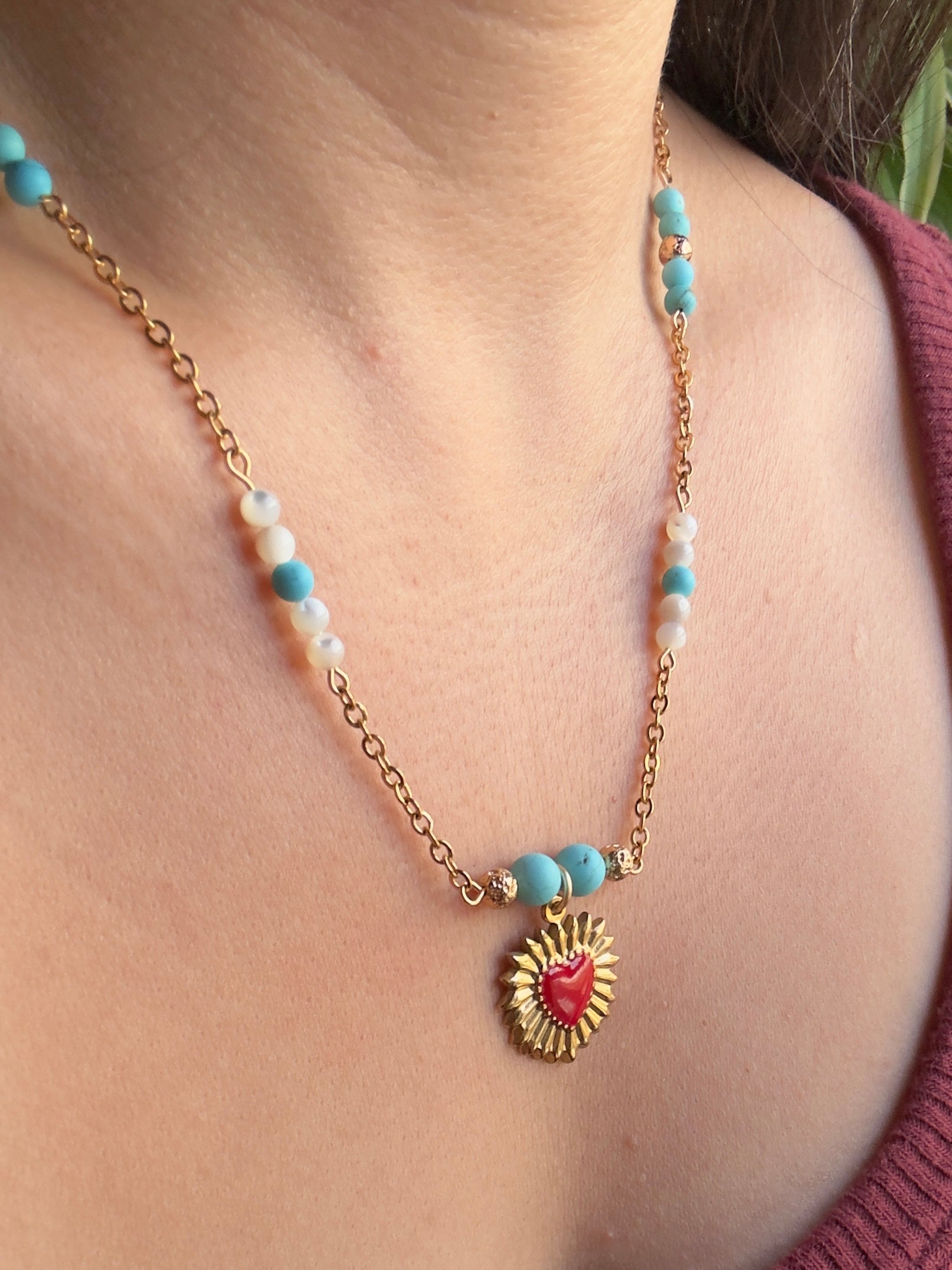 Red enamel gold stainless steel heart pendant on gold stainless steel chain with blue howlite and mother of pearl beads, slight side view close up on model