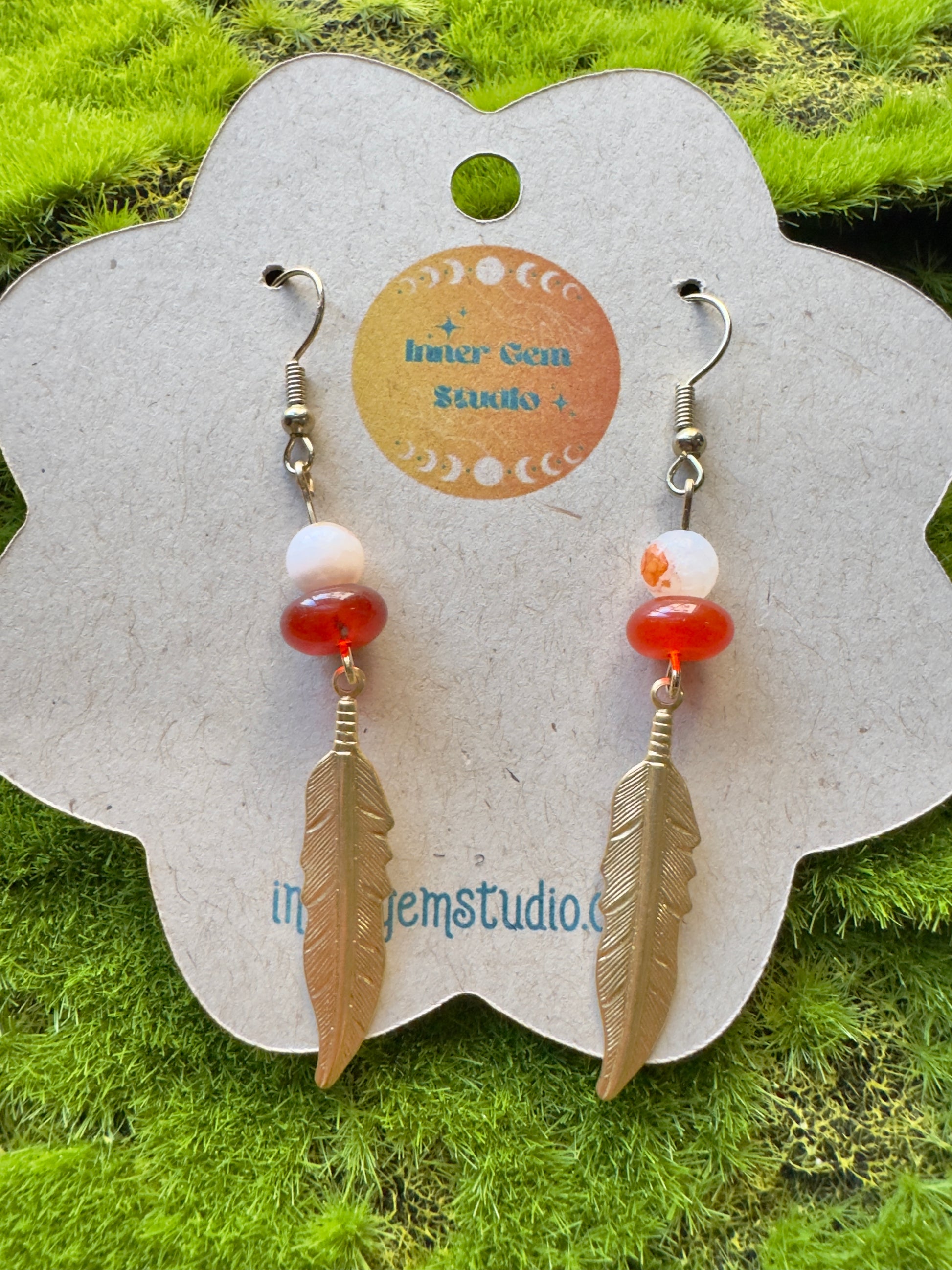 Feather-shaped earrings with red and white beads on a display card against a grassy background