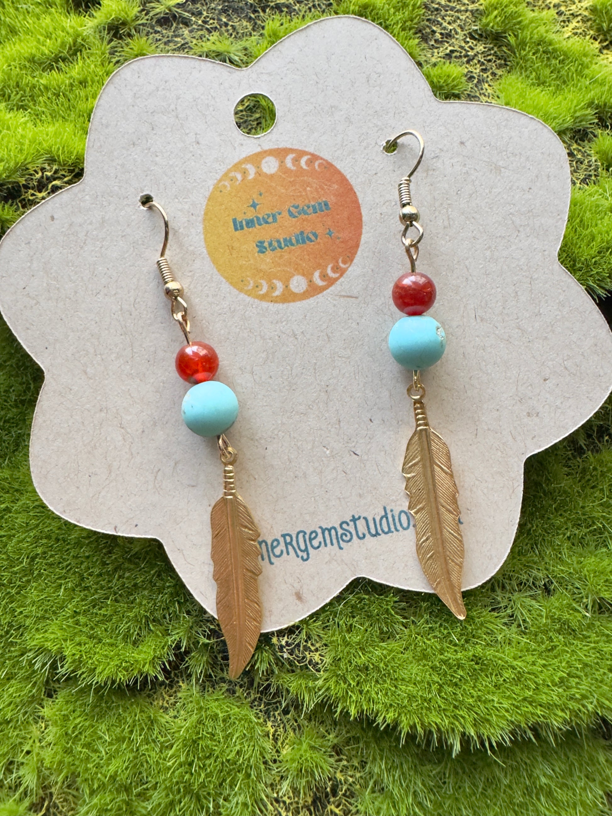 Dangling feather-shaped earrings with beads on a branded card against a grassy background