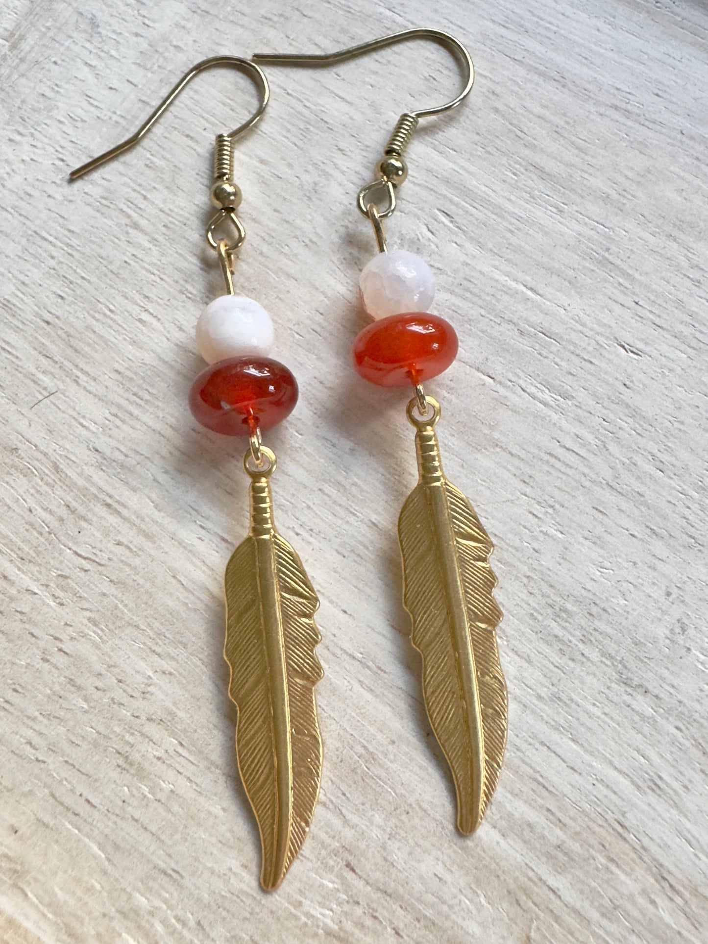 Gold feather earrings with red and white beads on a light wooden surface
