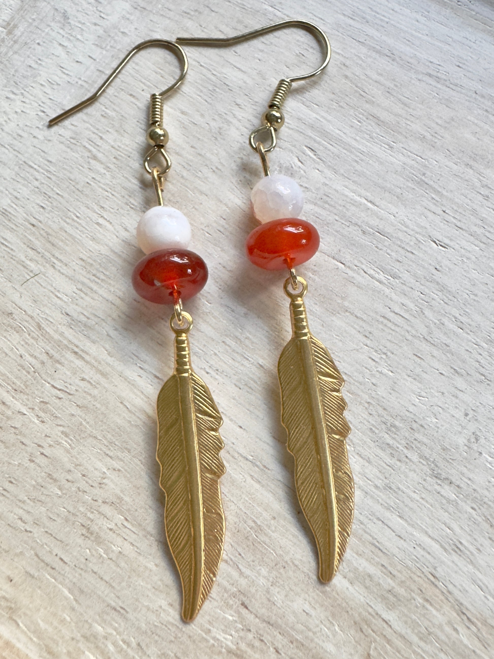 Gold feather earrings with red and white beads on a light wooden surface