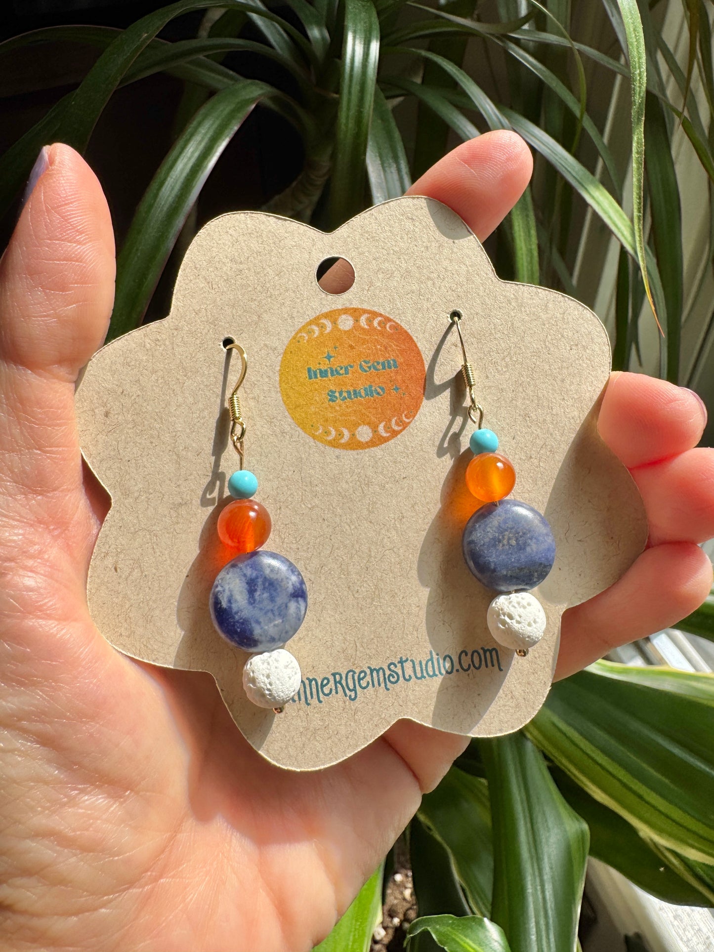 A hand holds a flower-shaped kraft display card featuring a pair of dangling earrings. The earrings have gold hooks, turquoise beads, orange beads, a blue stone bead, and white textured beads.
