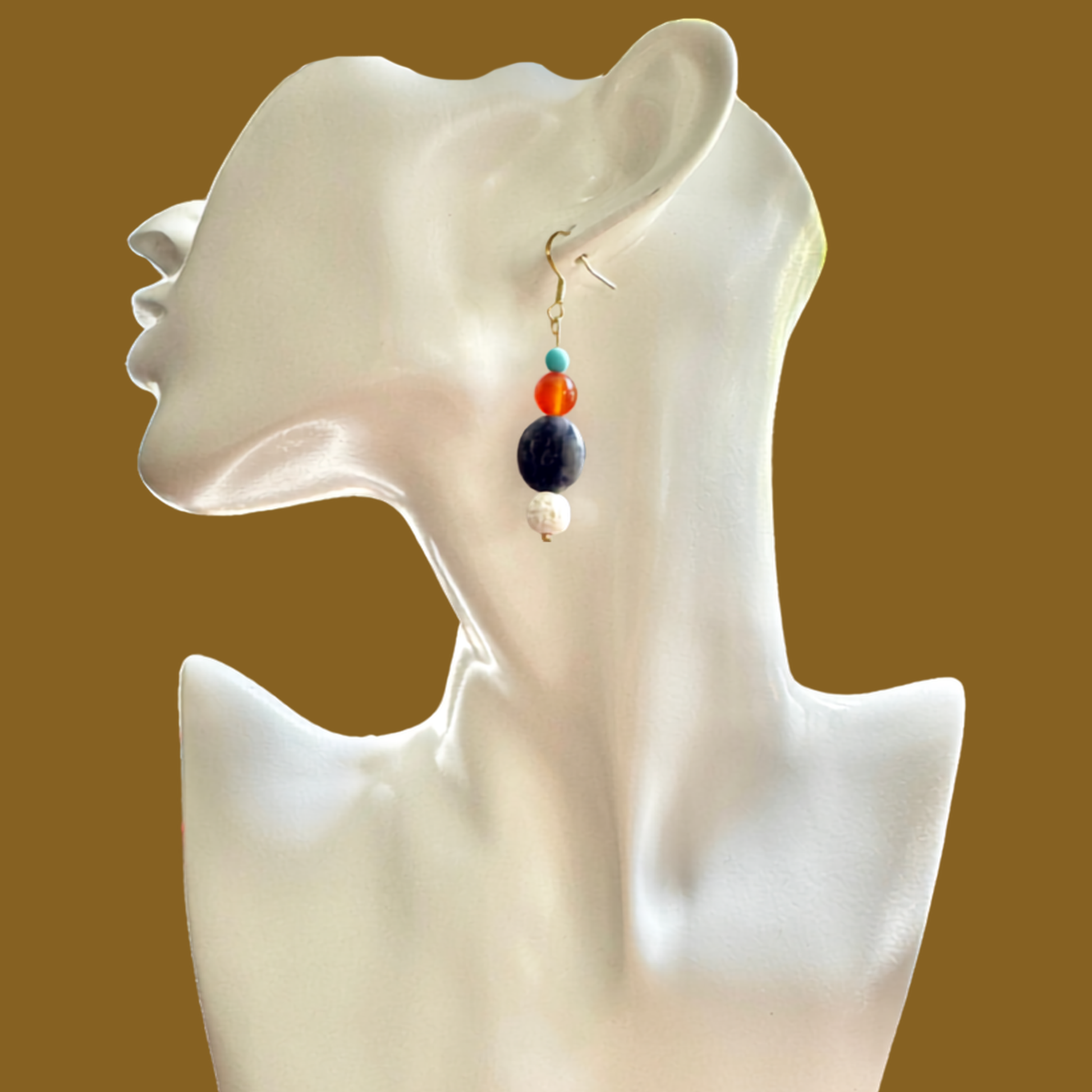 A single dangling beaded earring displayed on a glossy white female bust against a solid warm brown background. The earring features a gold fishhook clasp with stacked beads in turquoise, orange, dark navy blue, and white.