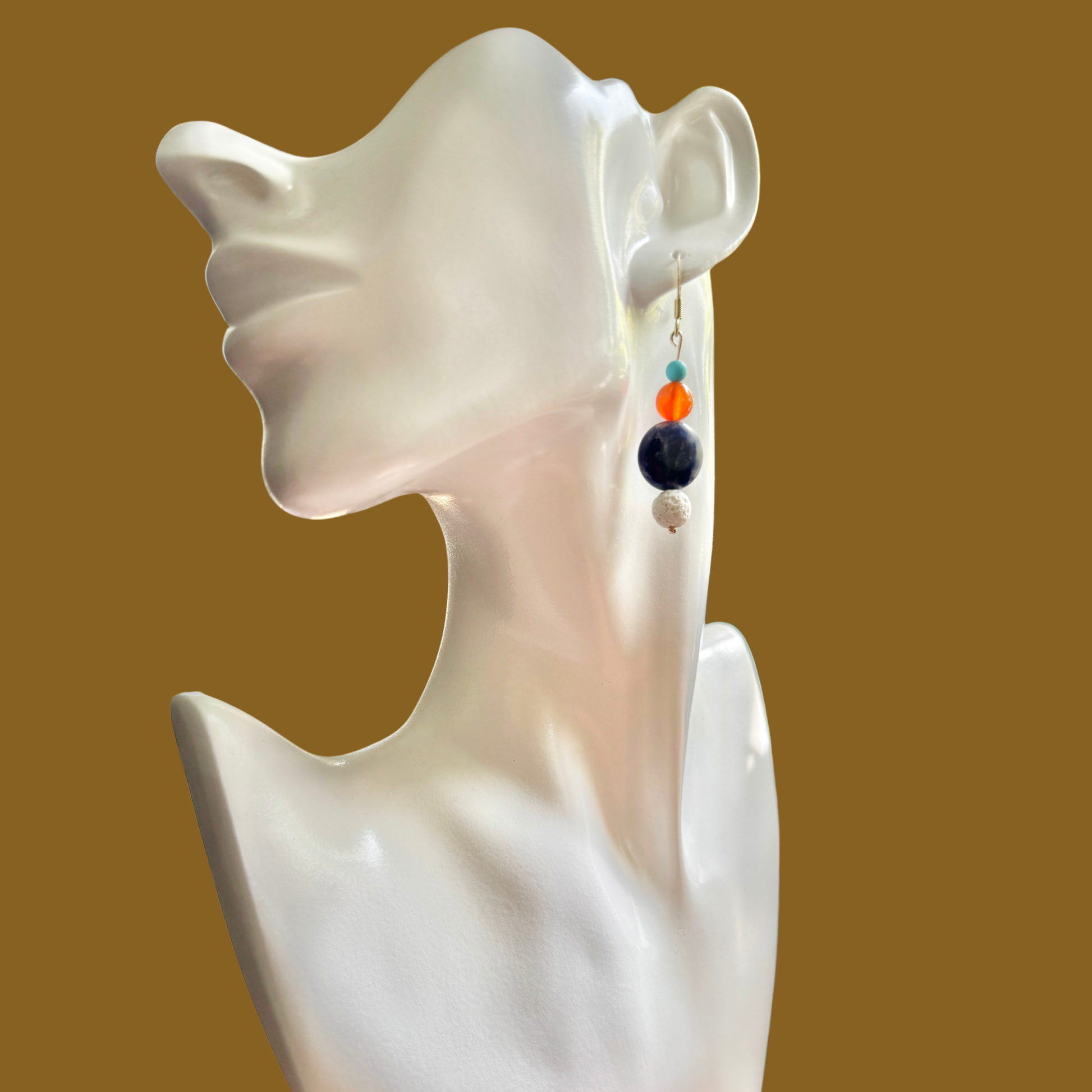 A single dangling beaded earring displayed on a glossy white ceramic female bust against a solid warm brown background. The earring features a silver-toned hook, a small turquoise bead, an orange bead, a dark navy blue bead, and a tiny clear bead at the bottom.