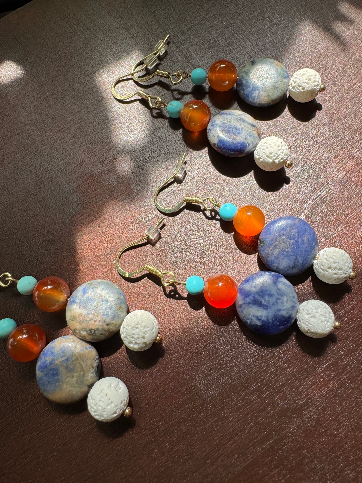 A pair of handmade beaded drop earrings laid on a dark wooden surface. The earrings feature large blue and white marbled beads, white textured beads, small turquoise beads, and orange agate beads, suspended from gold-toned fishhook wires.