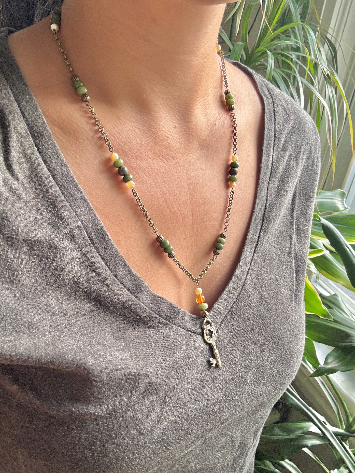 Necklace with green and orange beads and a key pendant worn by a person in a gray shirt, with plants in the background.