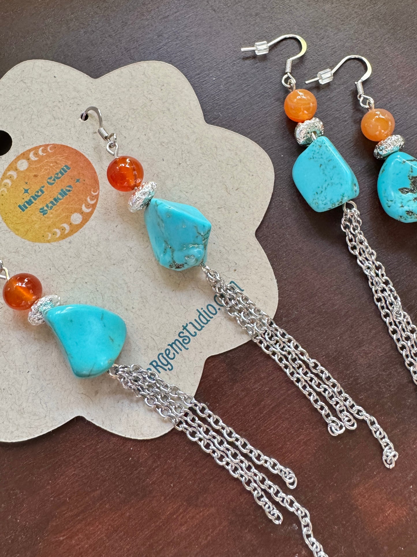 Boho-style dangle earrings with turquoise-colored stone pendants, orange beads, silver spacer rings, and silver chain drops, displayed on a kraft paper card on a dark wood surface.