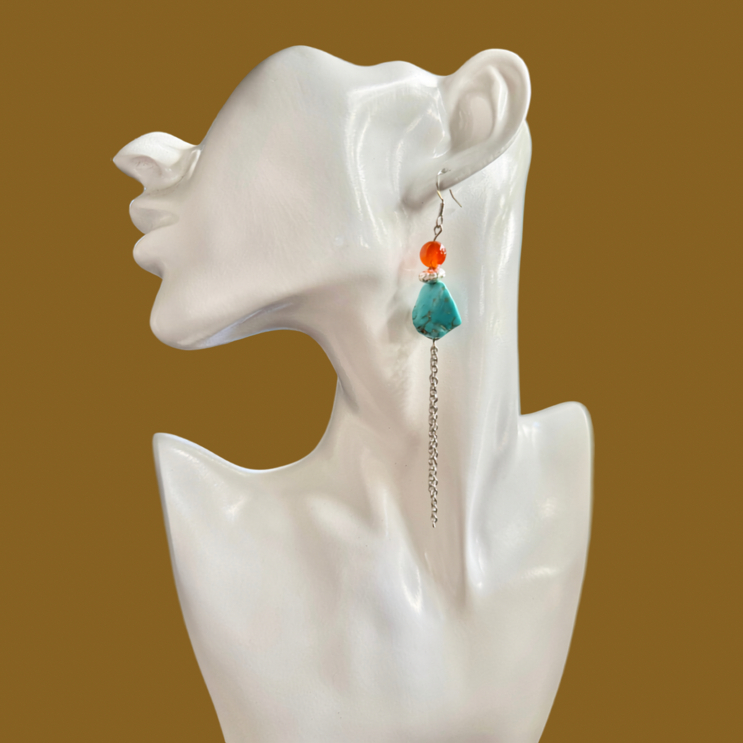 A single long dangle earring displayed on a white ceramic female bust against a solid warm brown background. The earring features a silver-toned chain, a small orange bead, and a larger teal-green teardrop-shaped stone.