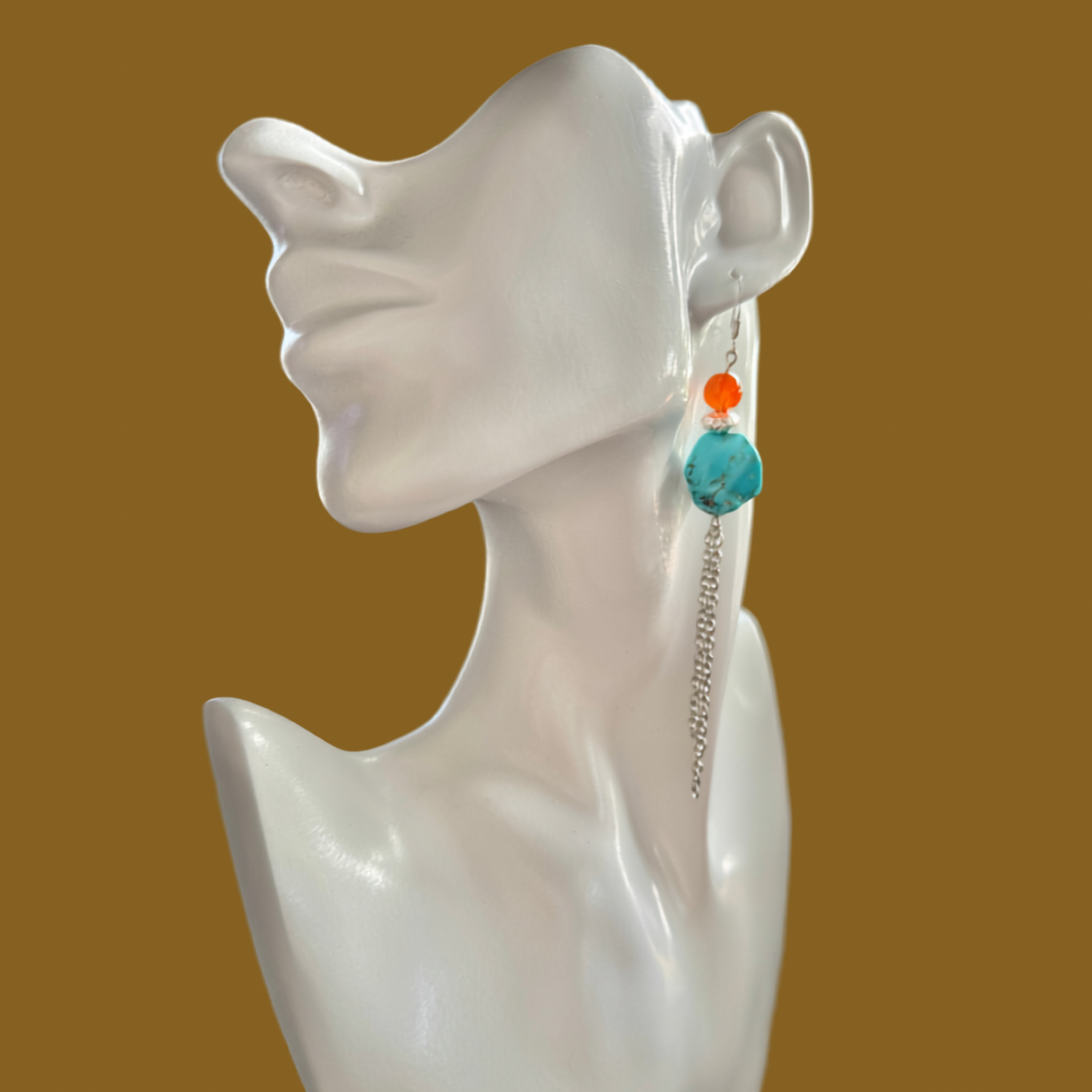 A pair of long dangle earrings displayed on a white female mannequin bust against a solid warm brown background. The earrings feature a silver-toned chain dangle with a round turquoise blue bead and a small orange bead accent.