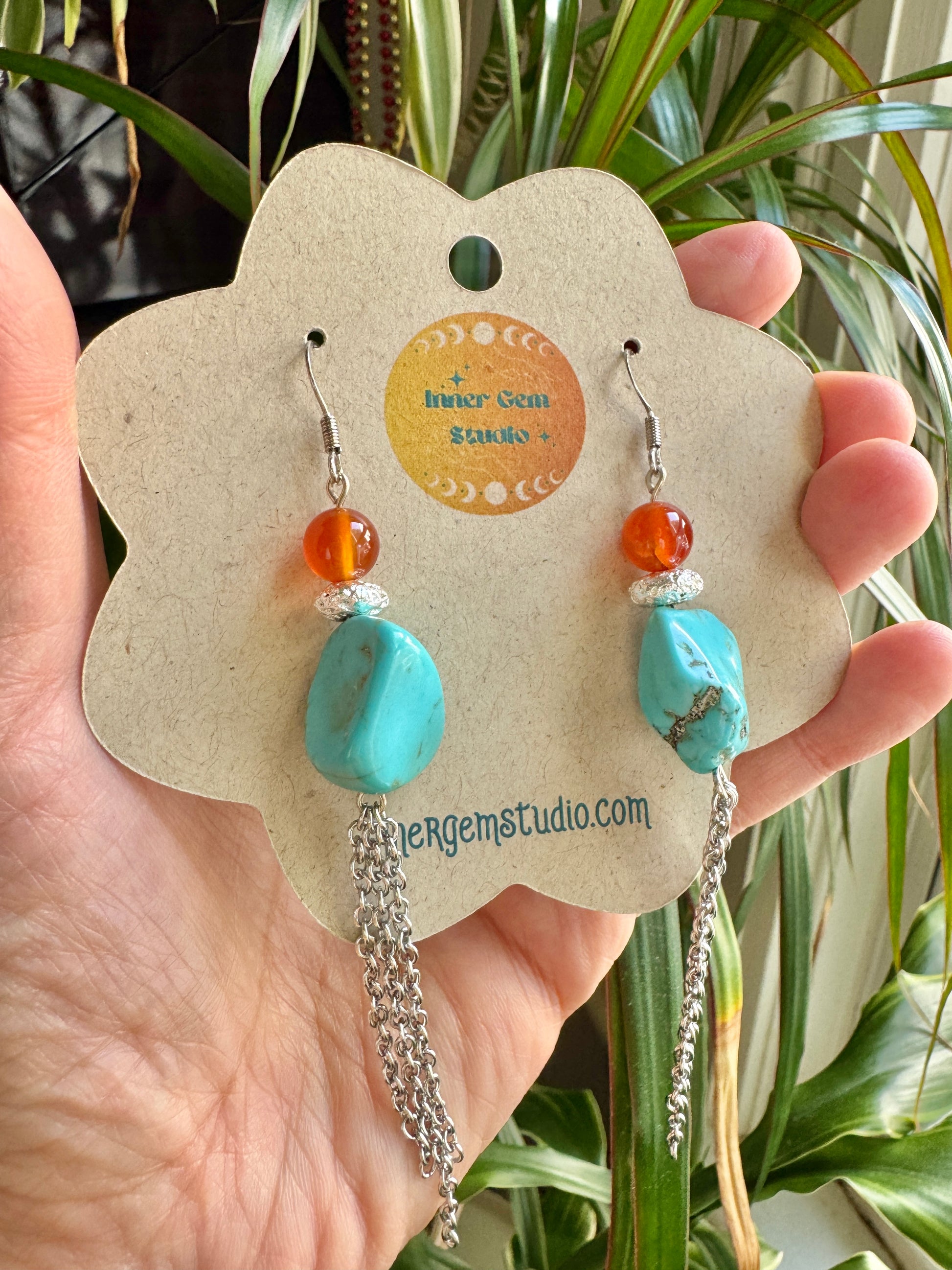 A hand holds a flower-shaped display card featuring a pair of dangling earrings. The earrings have turquoise-blue stone drops, small orange beads, silver spacer beads, and silver chain tassels.