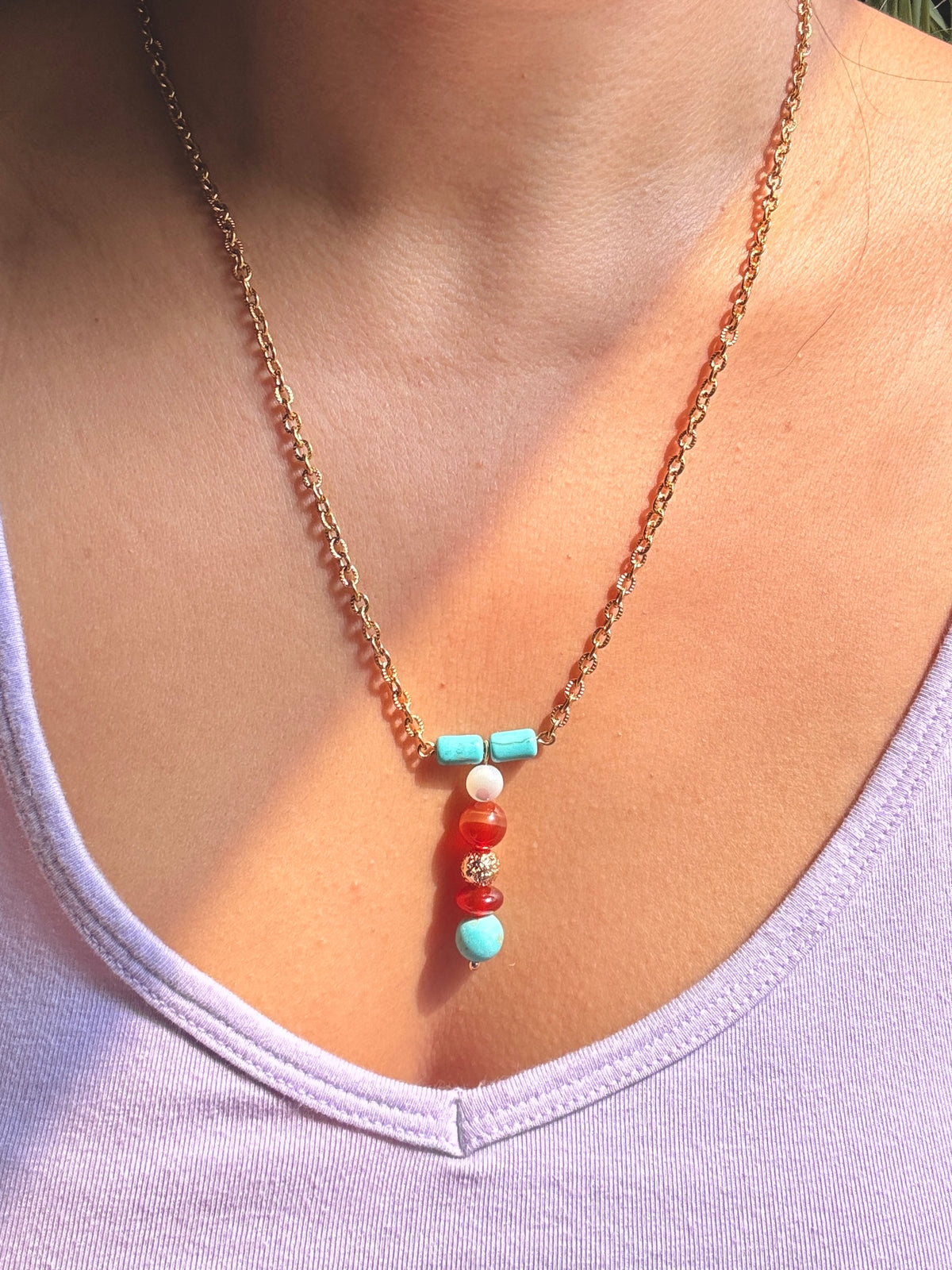 A close-up of a person wearing a delicate gold chain necklace with a beaded pendant. The pendant features turquoise blue beads, a white bead, red agate beads, and a small gold spacer, worn over a light lavender tank top.