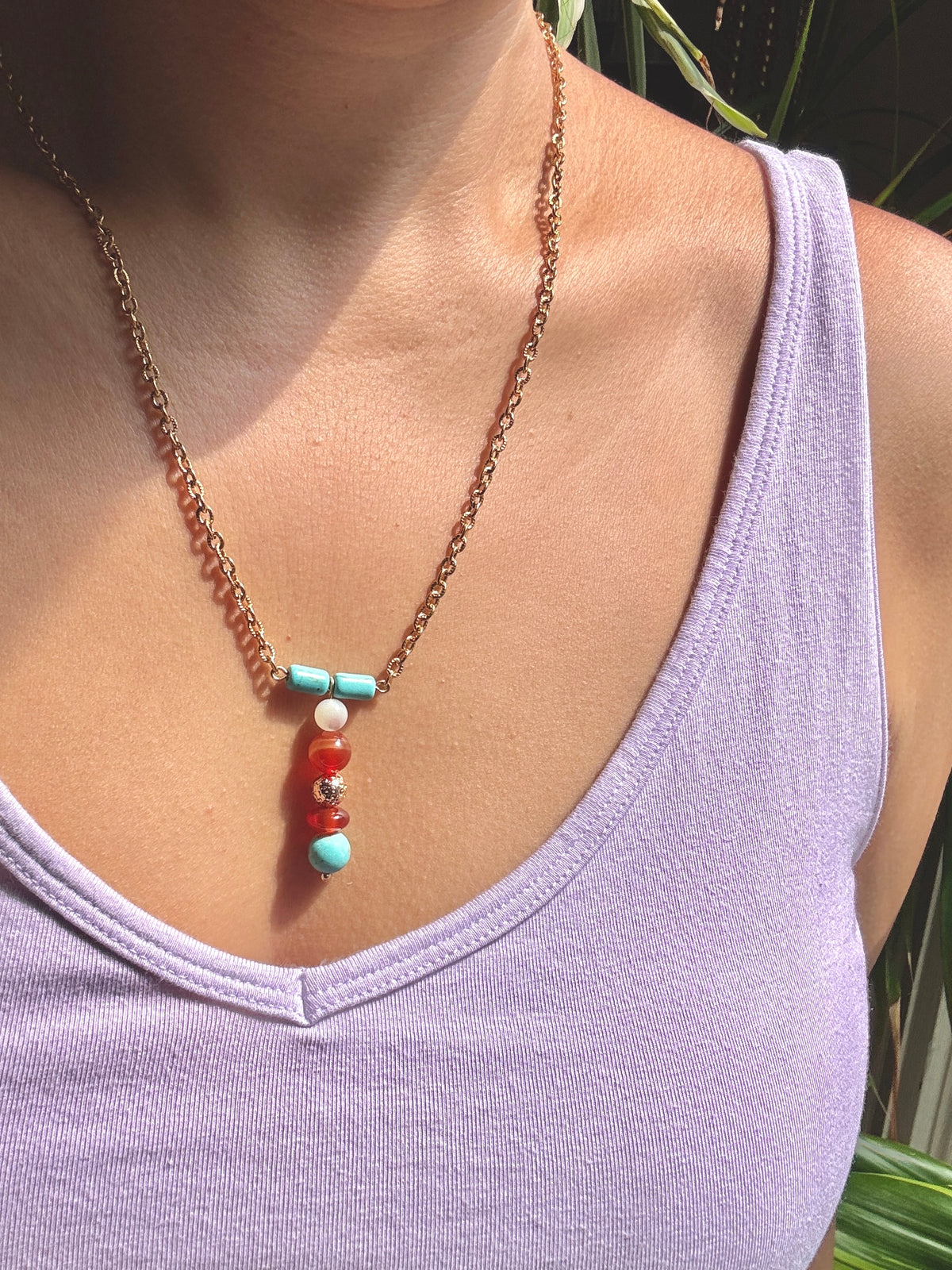 A close-up of a person wearing a delicate gold chain necklace with a vertical beaded pendant featuring turquoise, red, and white beads, paired with a light purple tank top.