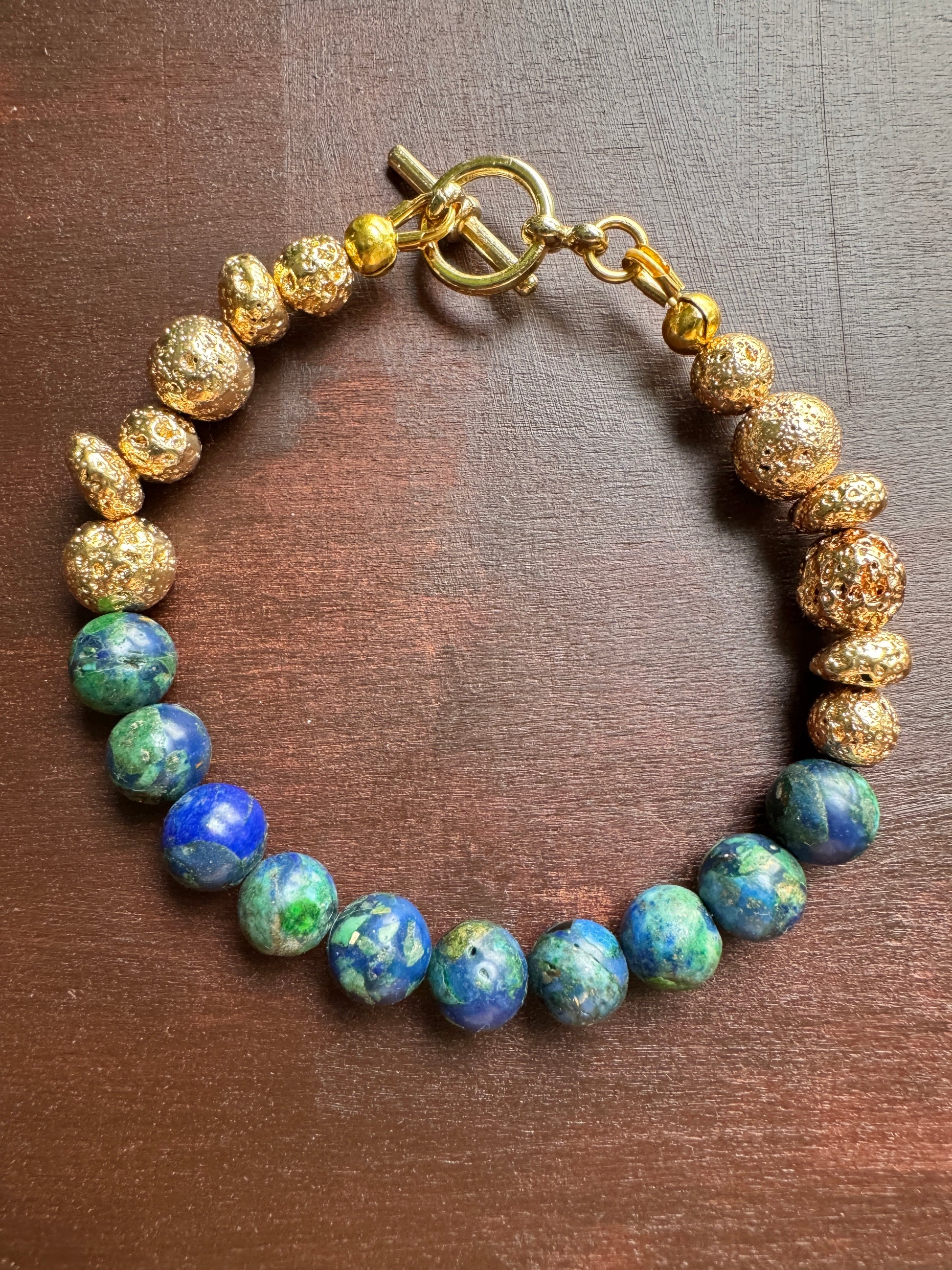 A beaded bracelet with textured gold-colored beads and round blue-green lapis lazuli-style beads, displayed on a dark brown wooden surface.