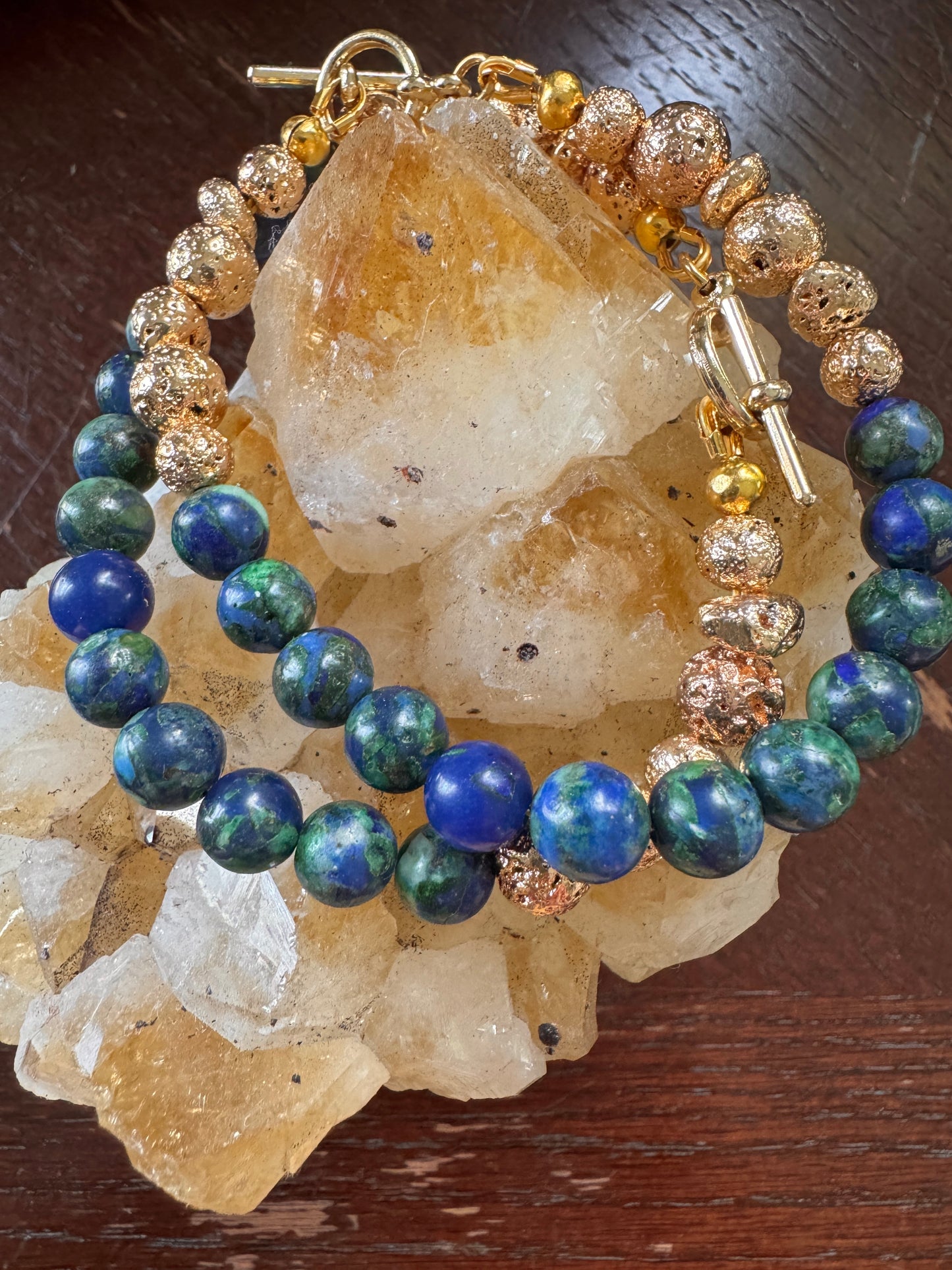 A beaded bracelet set displayed on a cluster of natural quartz crystals, featuring blue and green lapis lazuli beads, textured gold and rose gold metallic beads, and a gold toggle clasp, set against a dark wood background.
