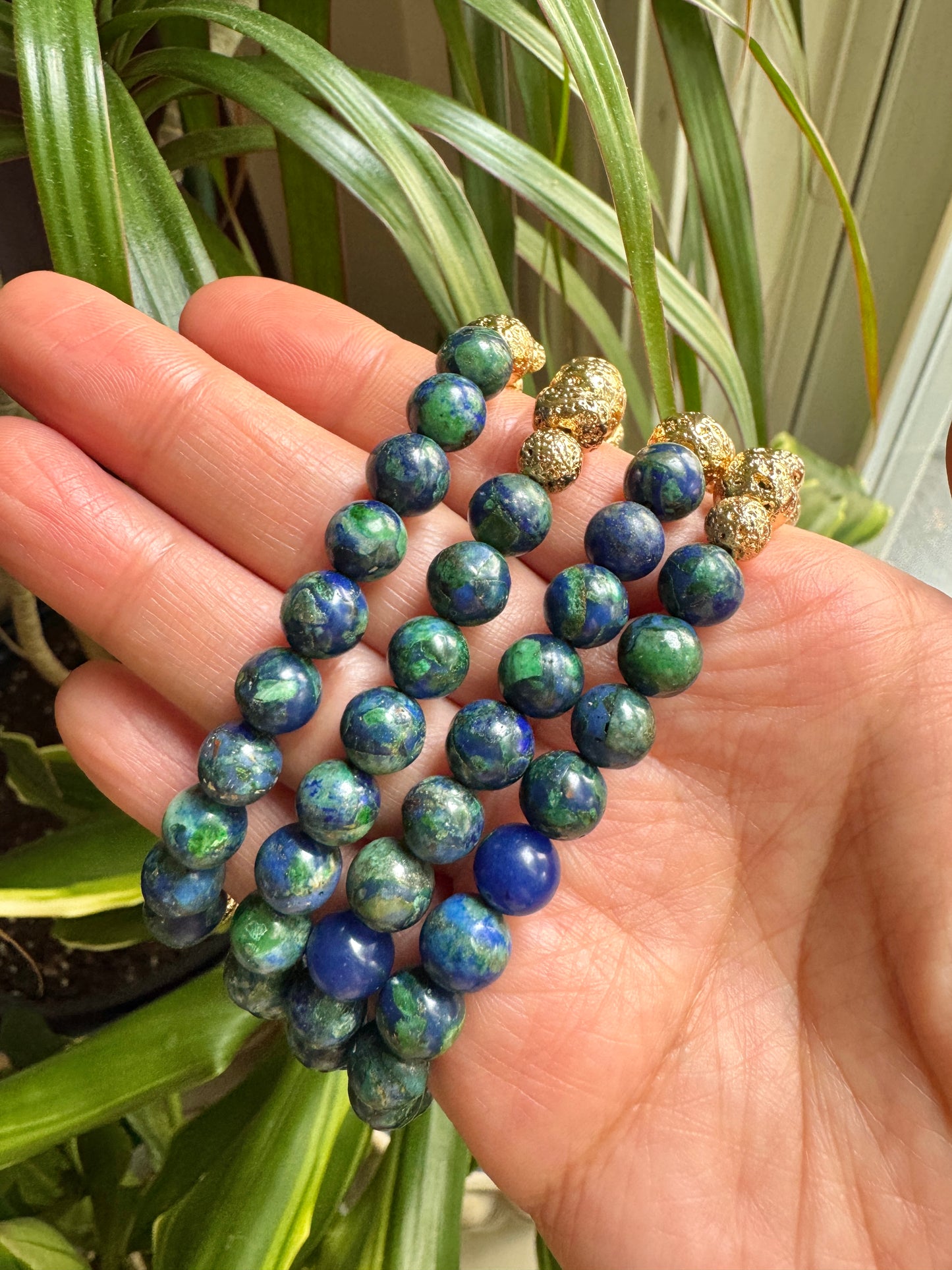 A hand holding three beaded bracelets. The bracelets feature round blue and green marbled beads, with one bracelet finished in textured gold-toned metal beads.