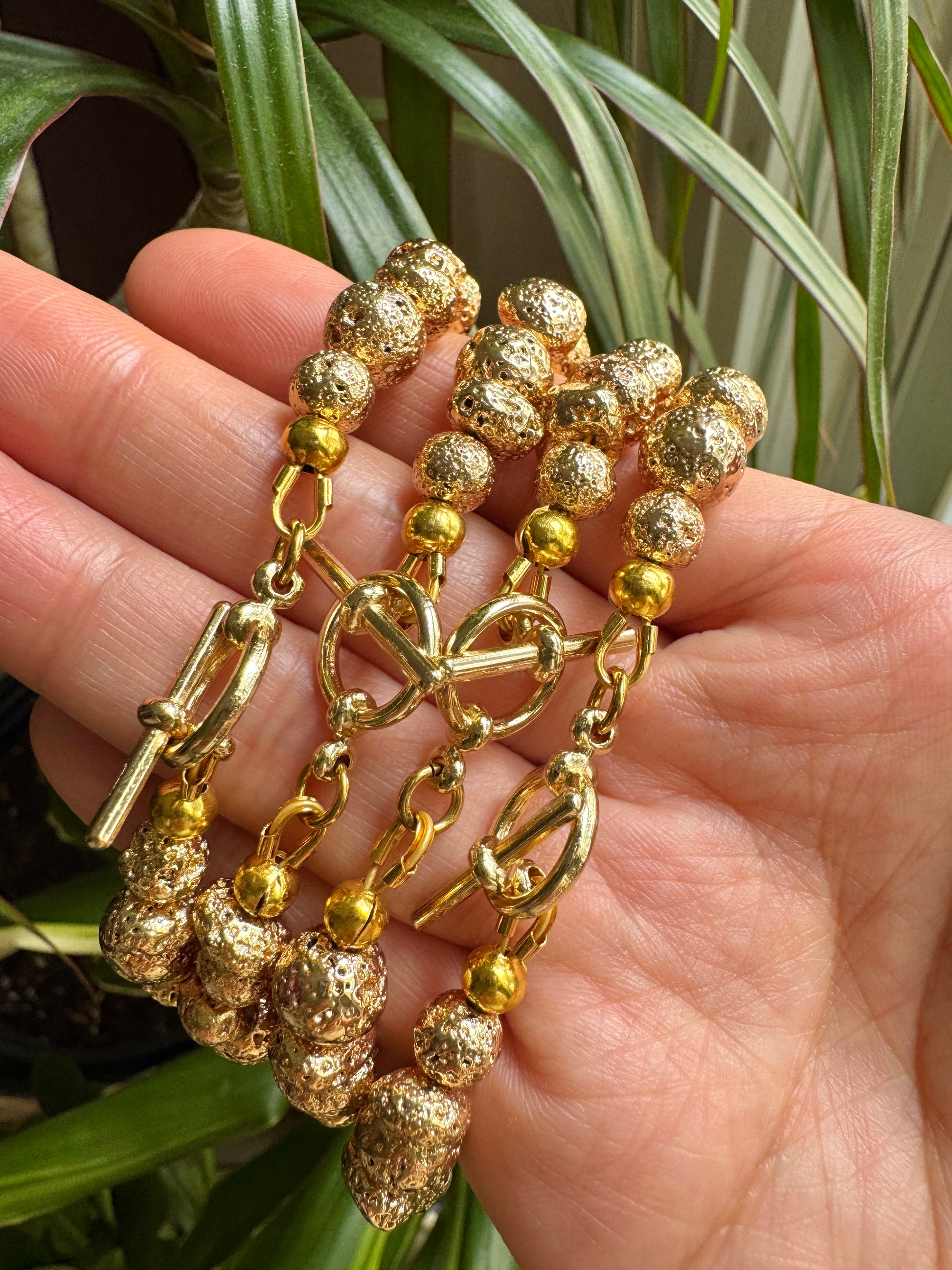 A gold-toned beaded bracelet with textured spherical beads, chain links, and a toggle clasp, held in a person's hand with green plant leaves in the background.