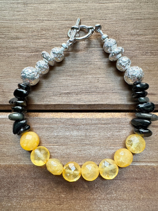 A beaded bracelet with faceted yellow beads, polished silver beads, and dark polished black beads, displayed on a wooden surface.