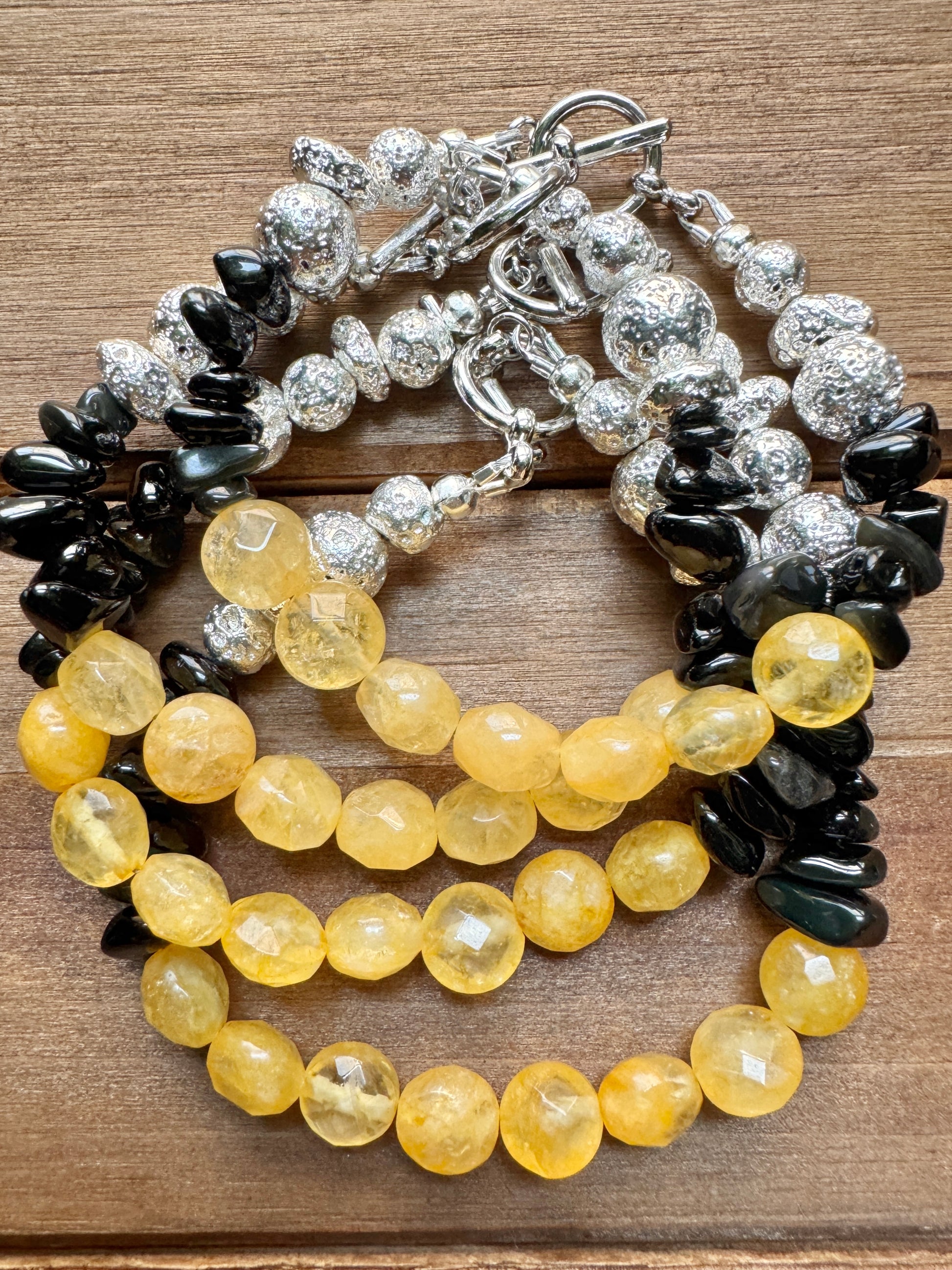 A multi-strand beaded bracelet featuring yellow faceted beads, black polished chips, and textured silver beads with a silver toggle clasp, displayed on a brown wooden surface.
