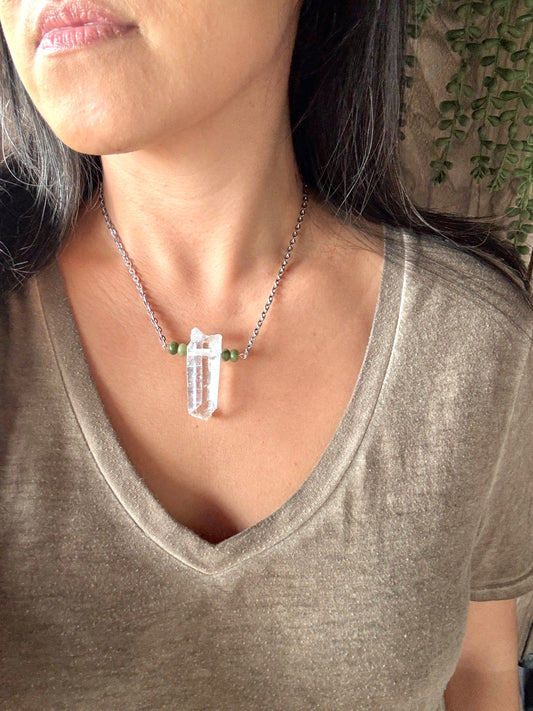 A close-up of a person wearing a silver chain necklace with a clear crystal pendant and small green beads on the chain, paired with a taupe V-neck top.
