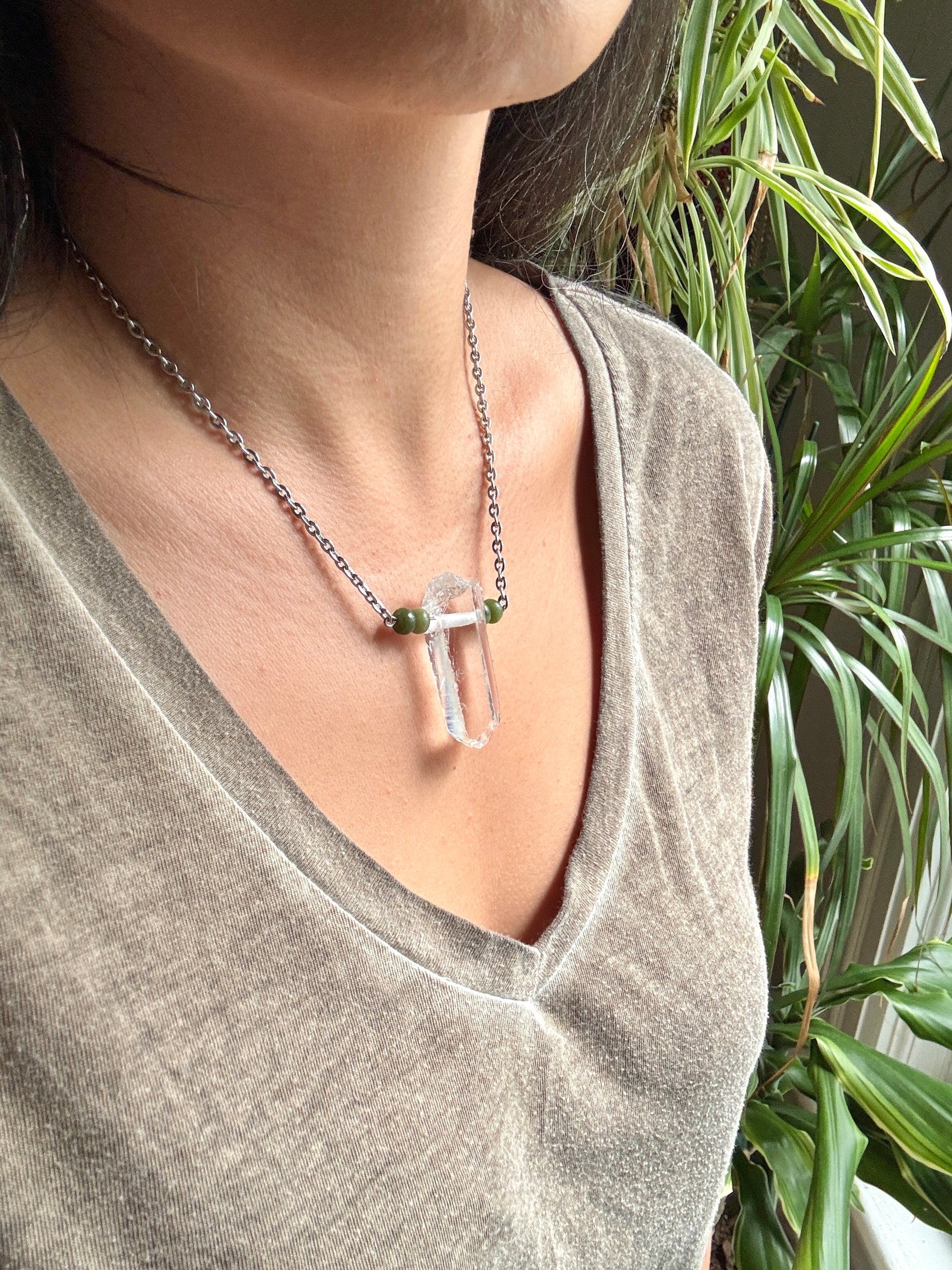 A close-up of a person wearing a silver-toned chain necklace with a clear crystal pendant and small green bead accents, paired with a heathered taupe-gray V-neck top, with green houseplants in the background.