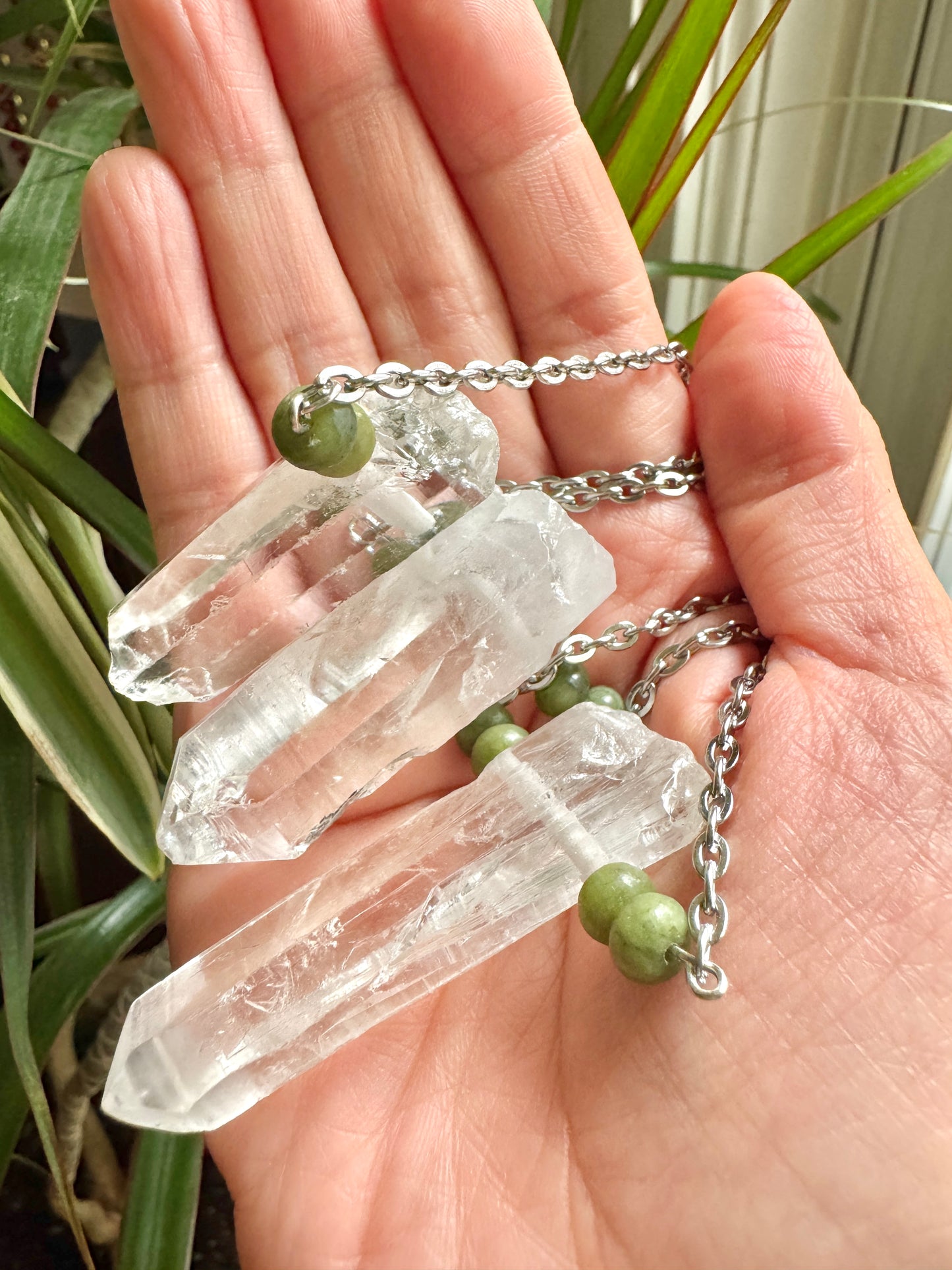 Three clear crystal points on silver chains with small green beads, held in a person's hand with green plants in the background.
