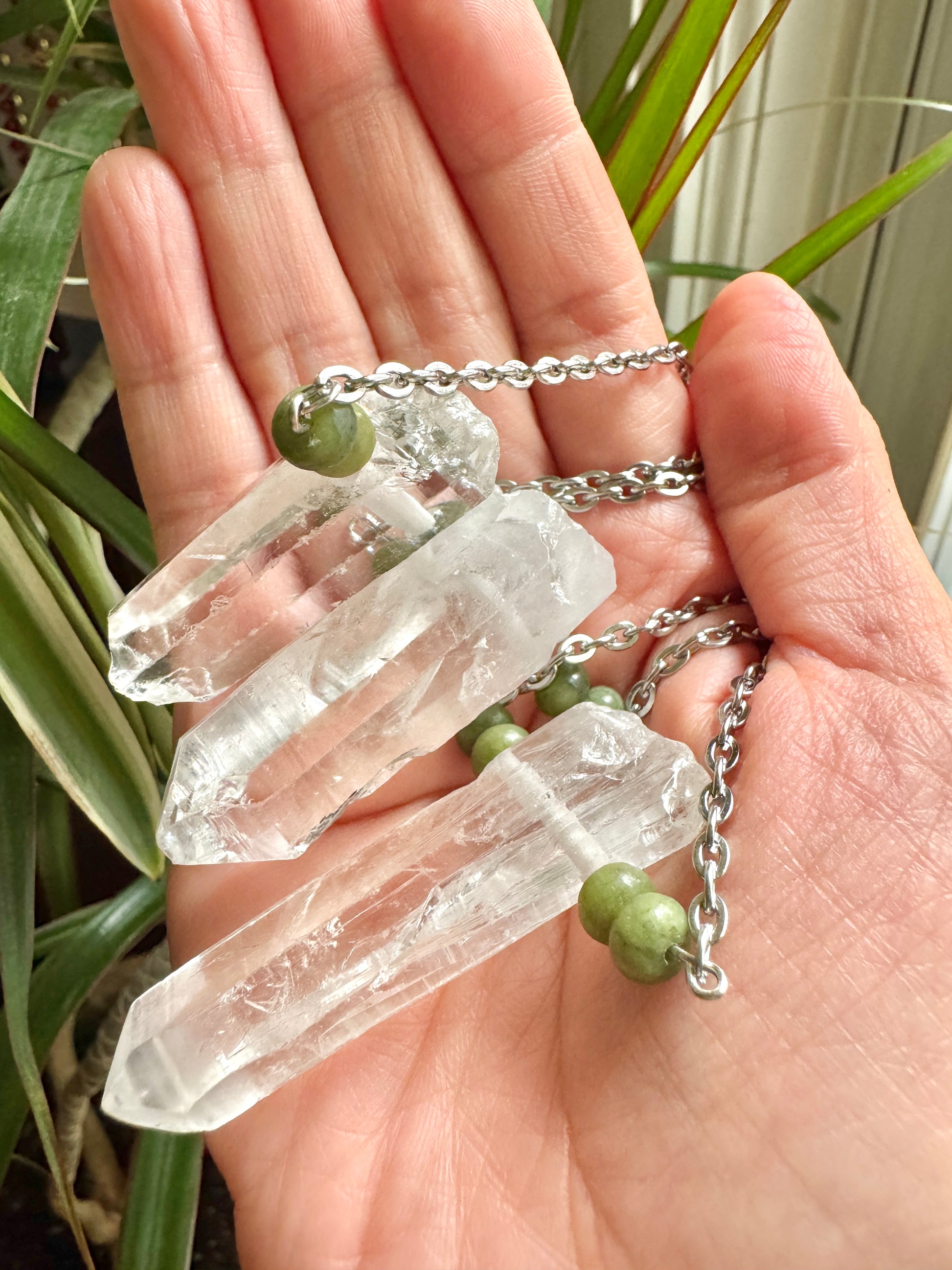 Three clear crystal points on silver chains with small green beads, held in a person's hand with green plants in the background.