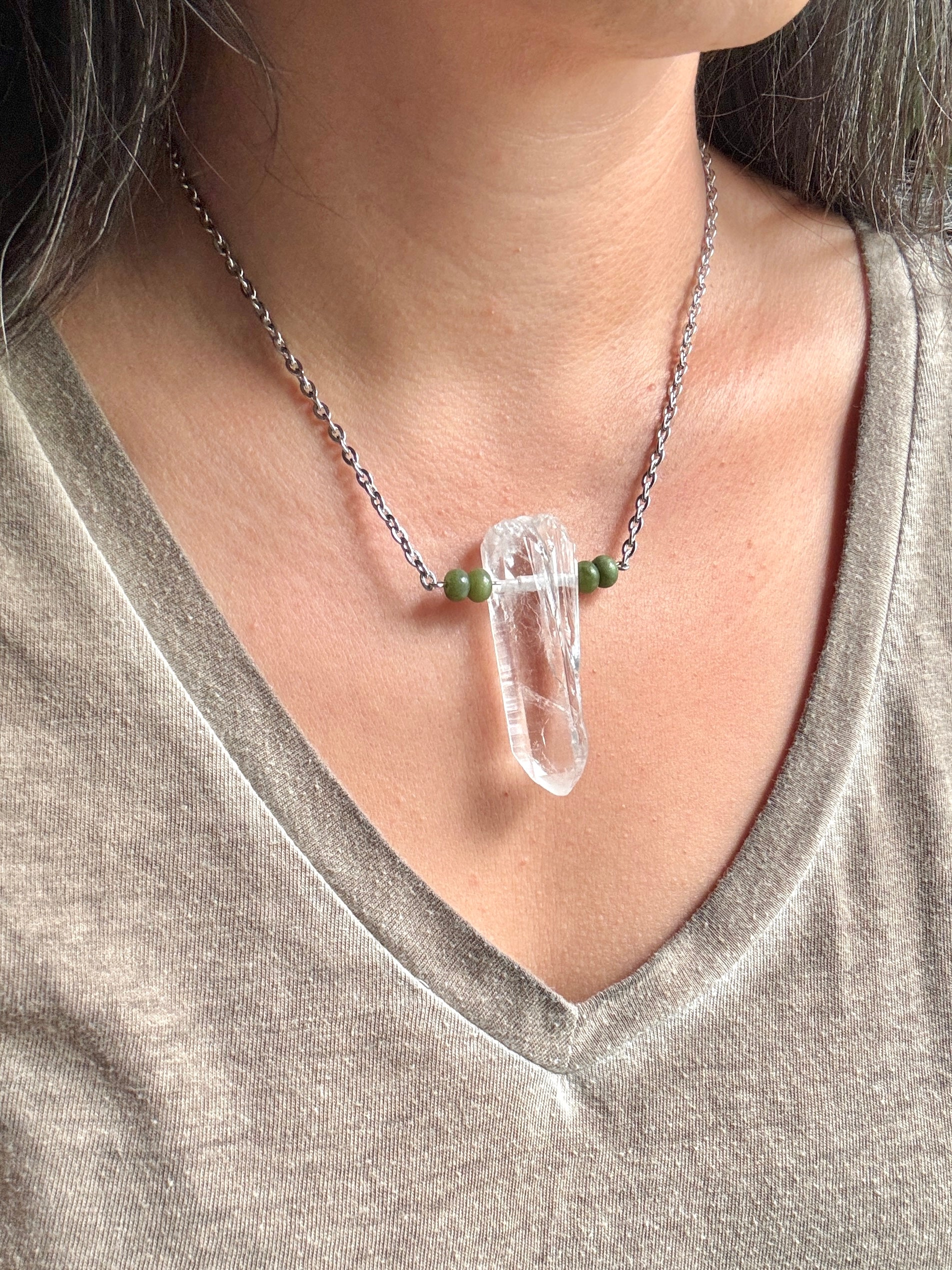 A close-up of a person wearing a silver chain necklace with a clear crystal point pendant and small green beads on either side, paired with a gray V-neck top.