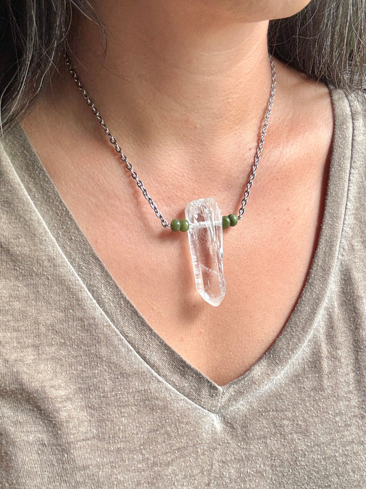 A close-up of a person wearing a silver chain necklace with a clear crystal point pendant and small green beads on either side, paired with a gray V-neck top.