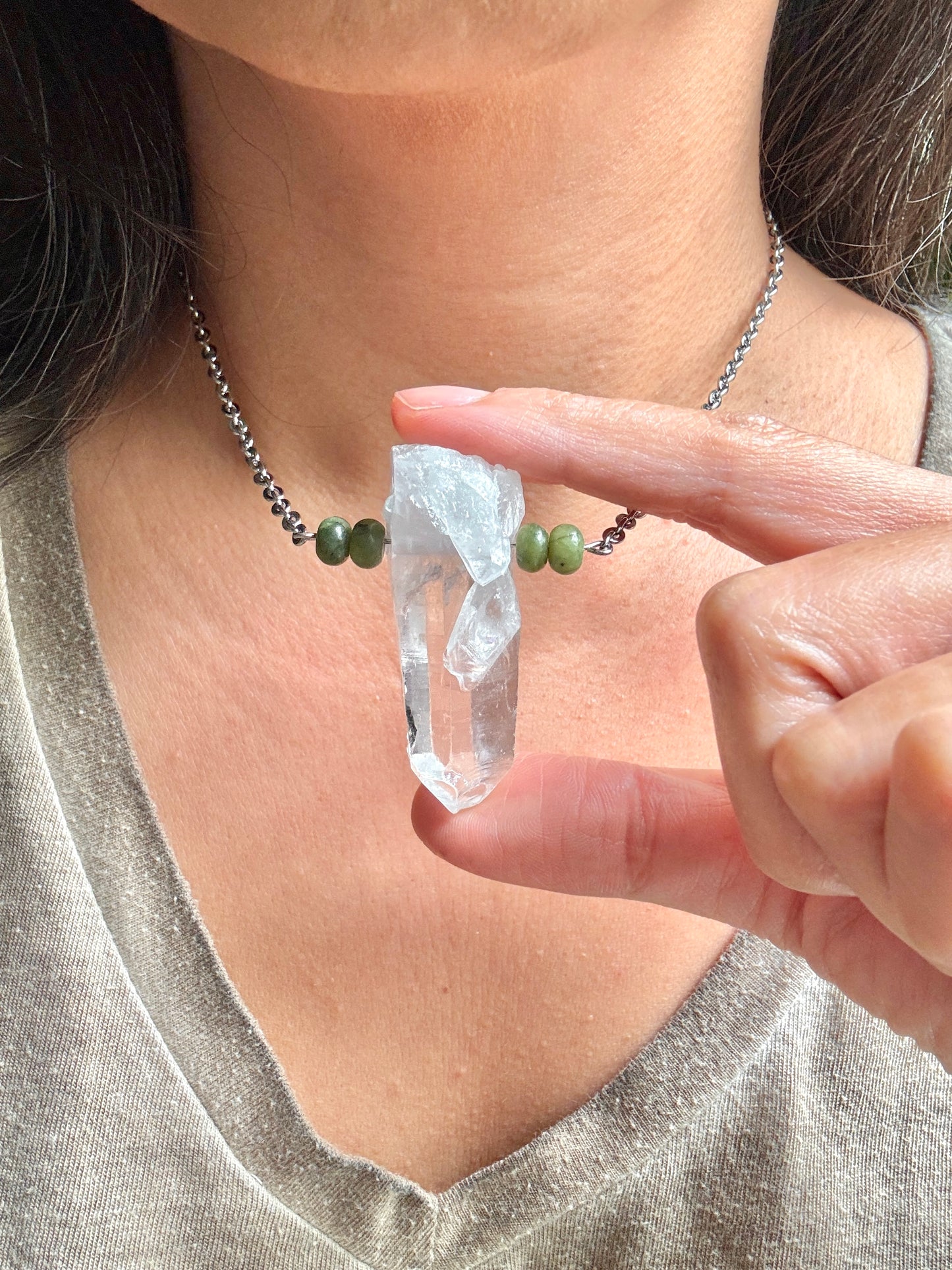 A close-up of a person wearing a silver beaded necklace with green beads and a clear pointed crystal pendant, while holding the pendant between their fingers.