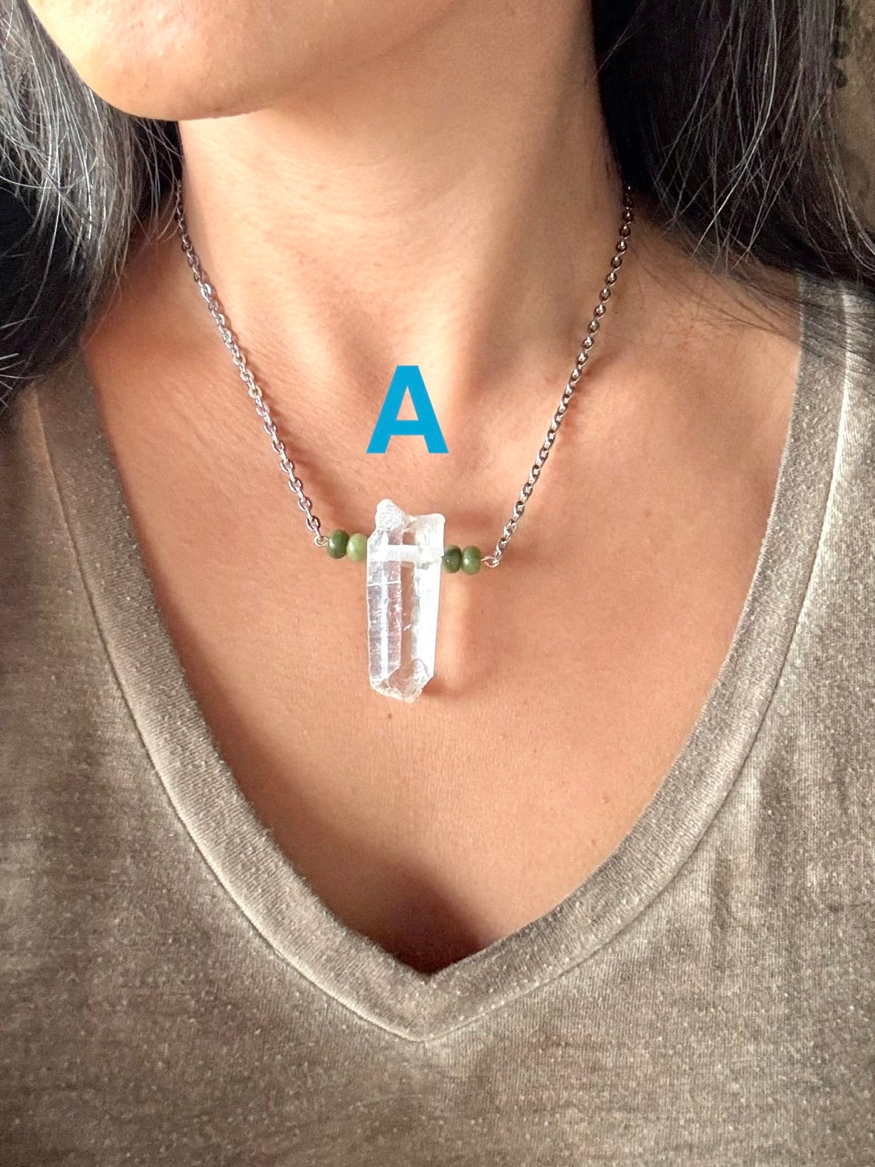 A close-up of a person's neck and chest wearing a silver chain necklace with a clear crystal pendant and small green beads, labeled with a blue letter 'A'.