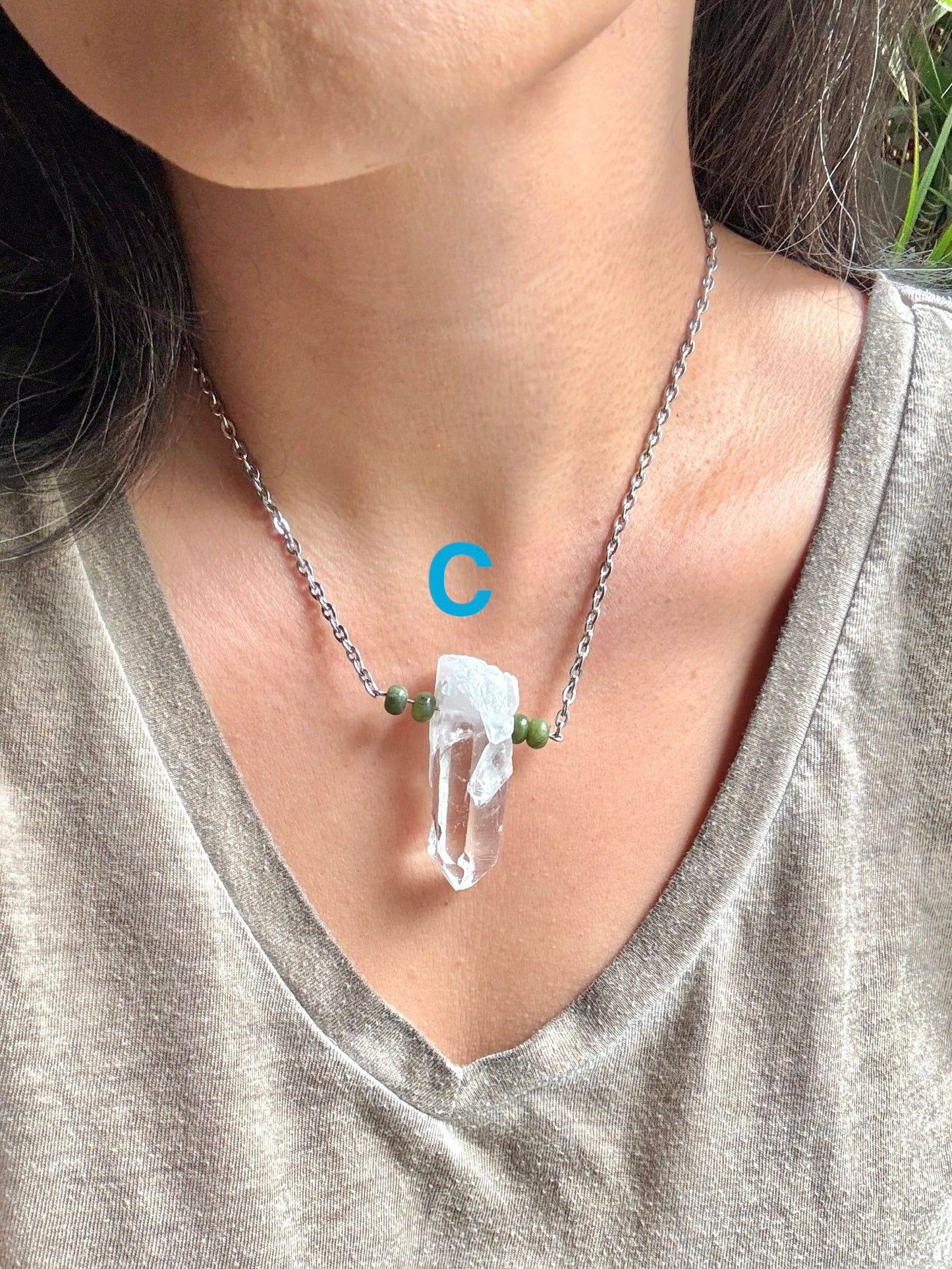 A close-up of a person wearing a silver chain necklace with a clear quartz crystal pendant and small green beads, paired with a gray v-neck top.