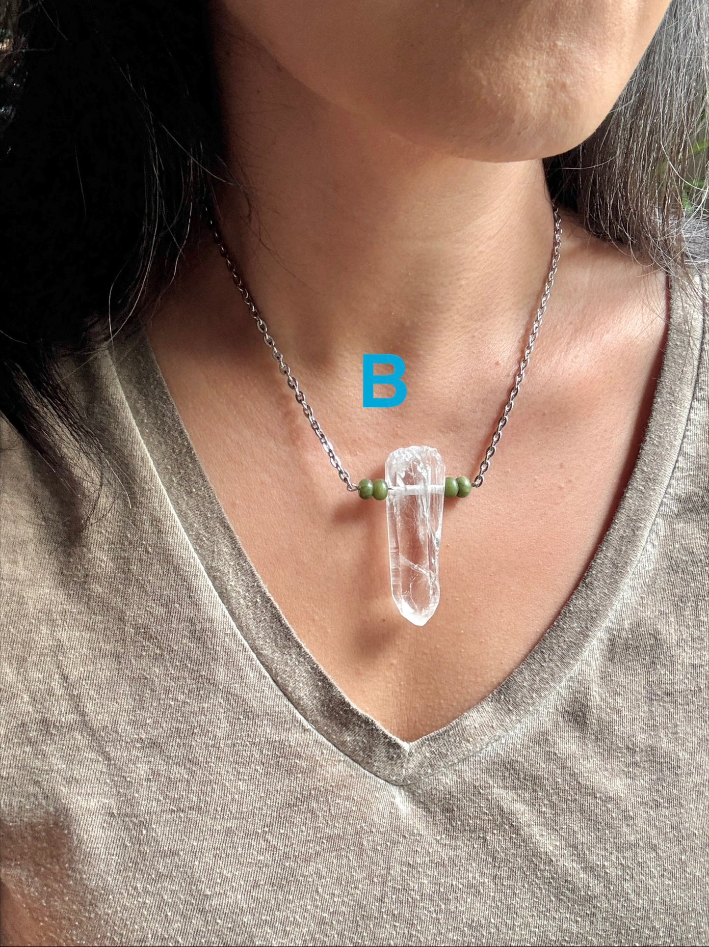 A close-up of a person wearing a silver chain necklace with a clear quartz crystal pendant and small green beads, shown against a gray V-neck top.