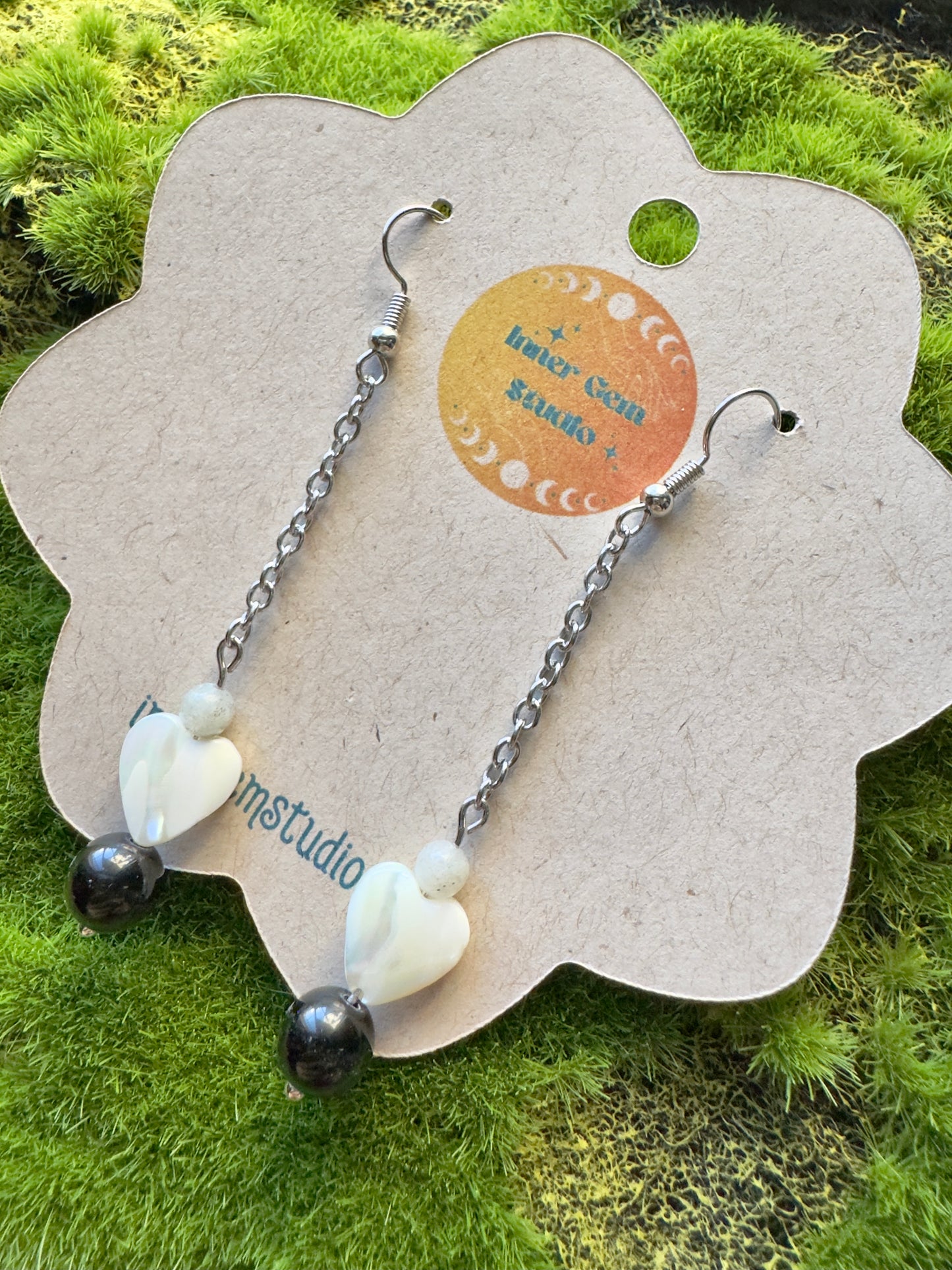 Pair of earrings with heart-shaped beads on a branded card against a grassy background