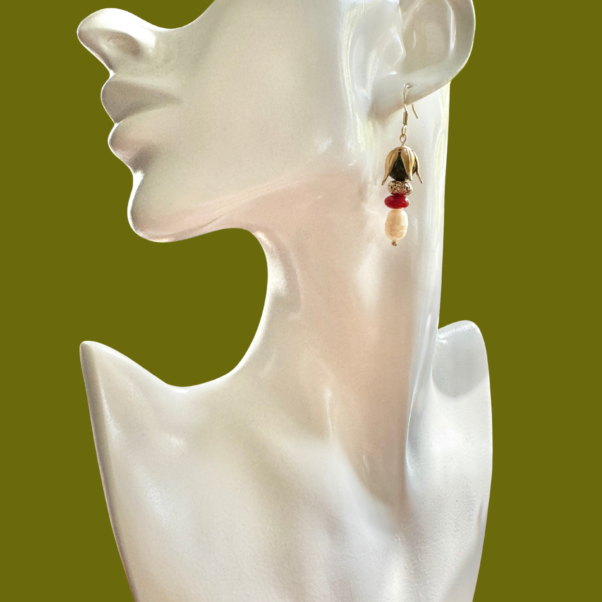 A dangling earring displayed on a glossy white female bust against a solid olive green background. The earring features a gold-toned top section, a small red bead, and a cream-colored bead at the bottom.