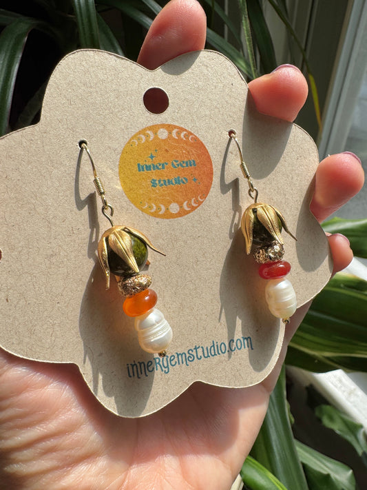 A hand holding a flower-shaped display card featuring a pair of dangling earrings. The earrings have gold-toned metal hooks and caps, with beads in olive green, orange, red, and white.