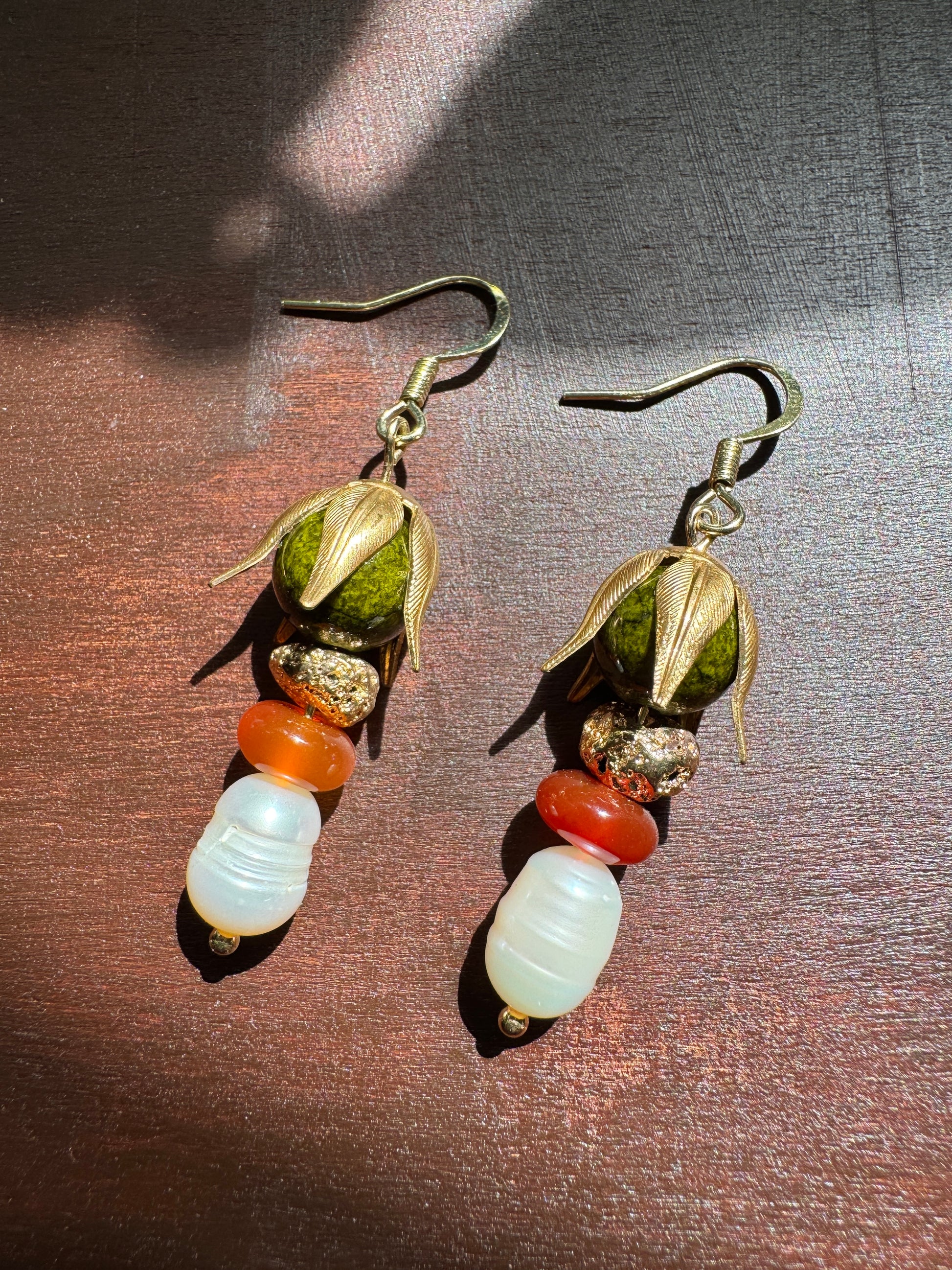 A pair of handmade dangle earrings featuring olive green seed pods, textured gold-toned beads, a translucent orange bead, and a large white ribbed bead at the bottom, displayed on a dark brown wooden surface.