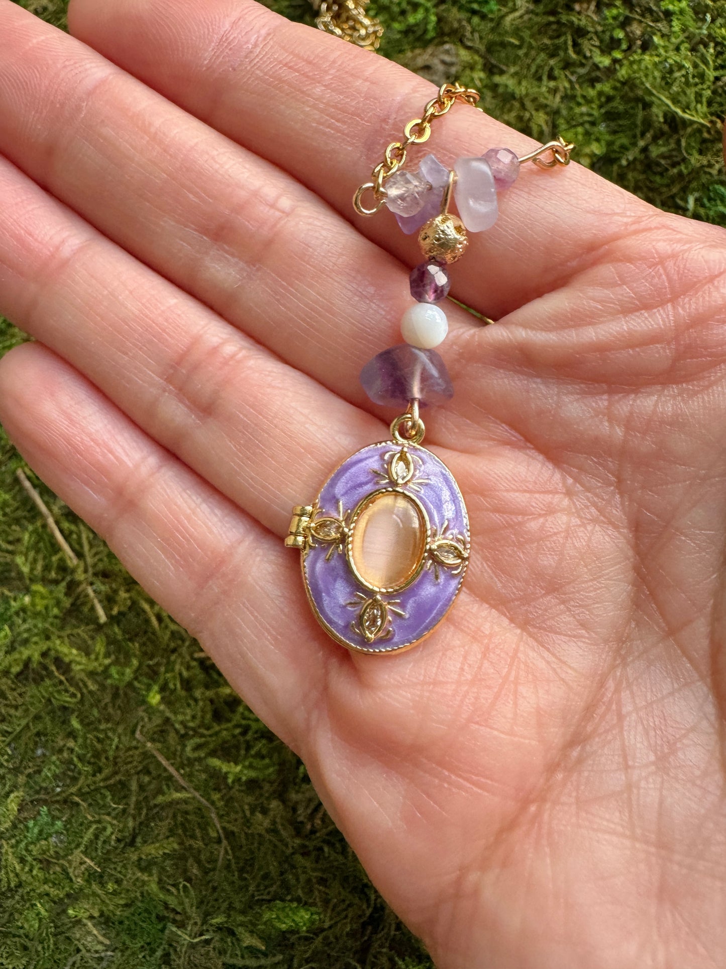 Hand holding a delicate necklace with a purple pendant against a green moss background