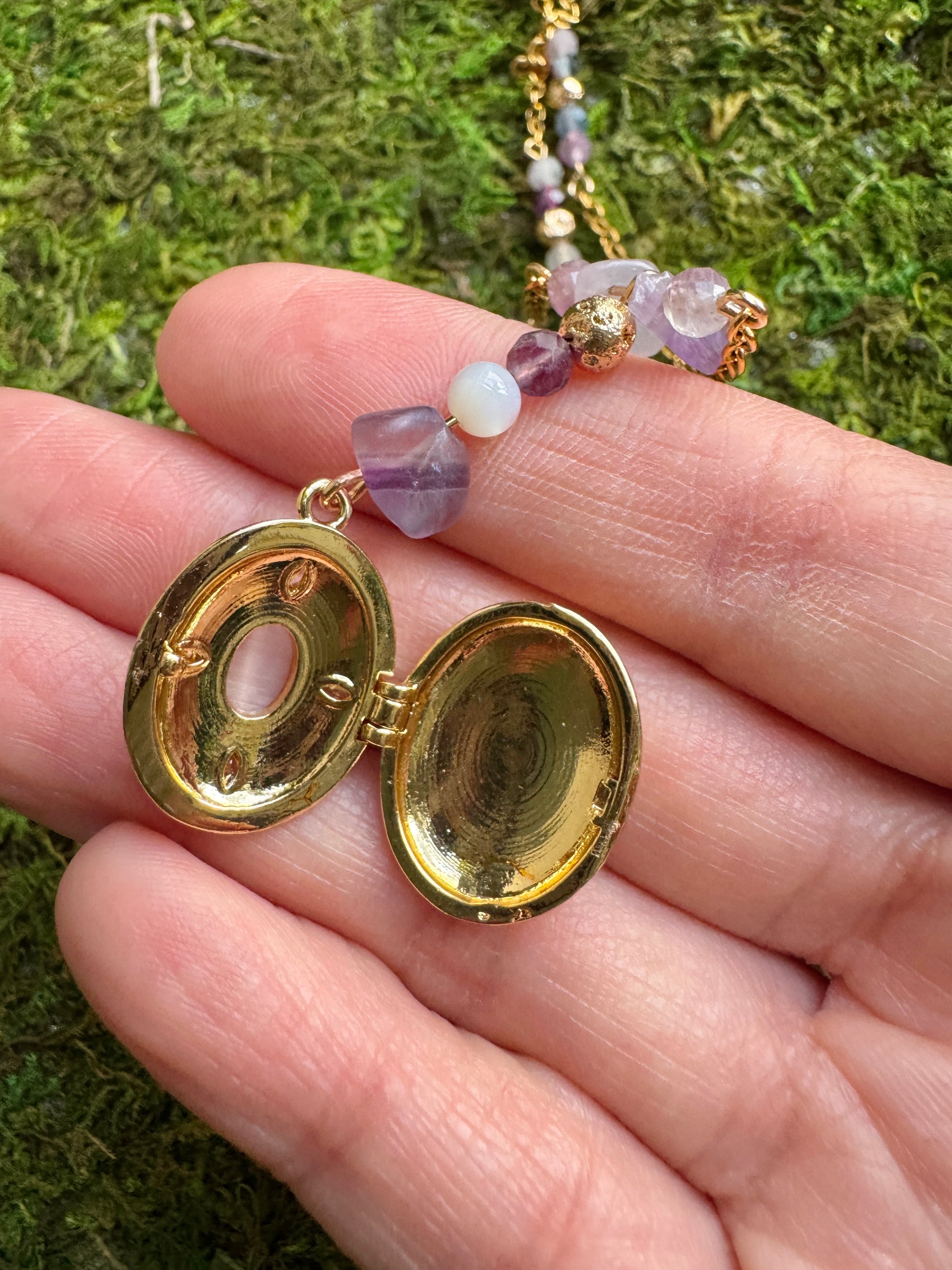 Gold locket with purple and white stones held in a hand against a grassy background
