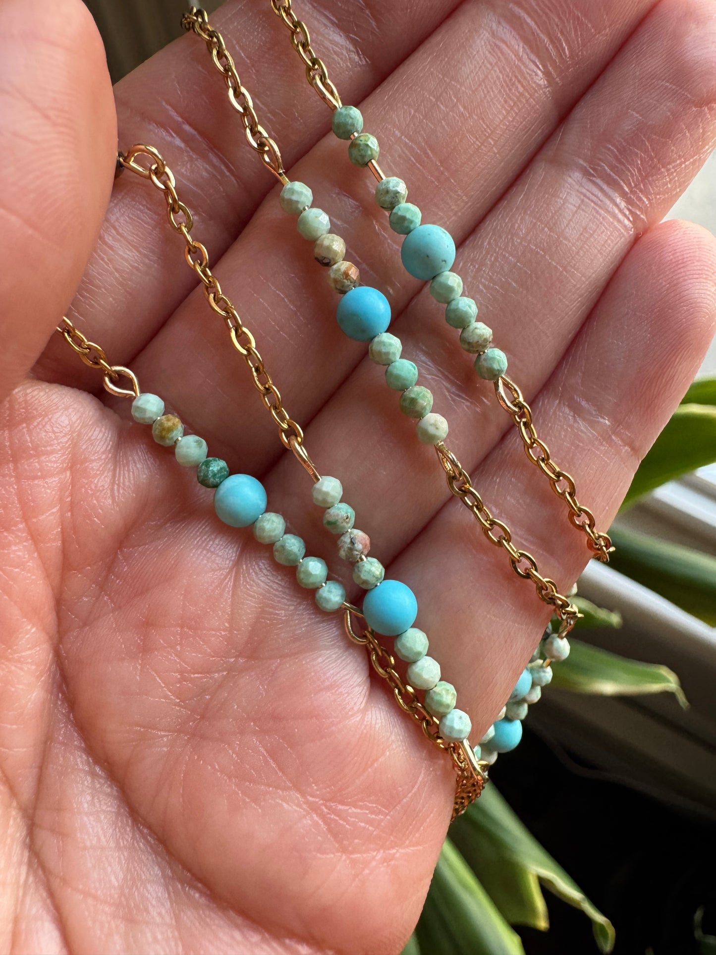Two beaded necklaces with gold chains held in a hand against a blurred natural background.