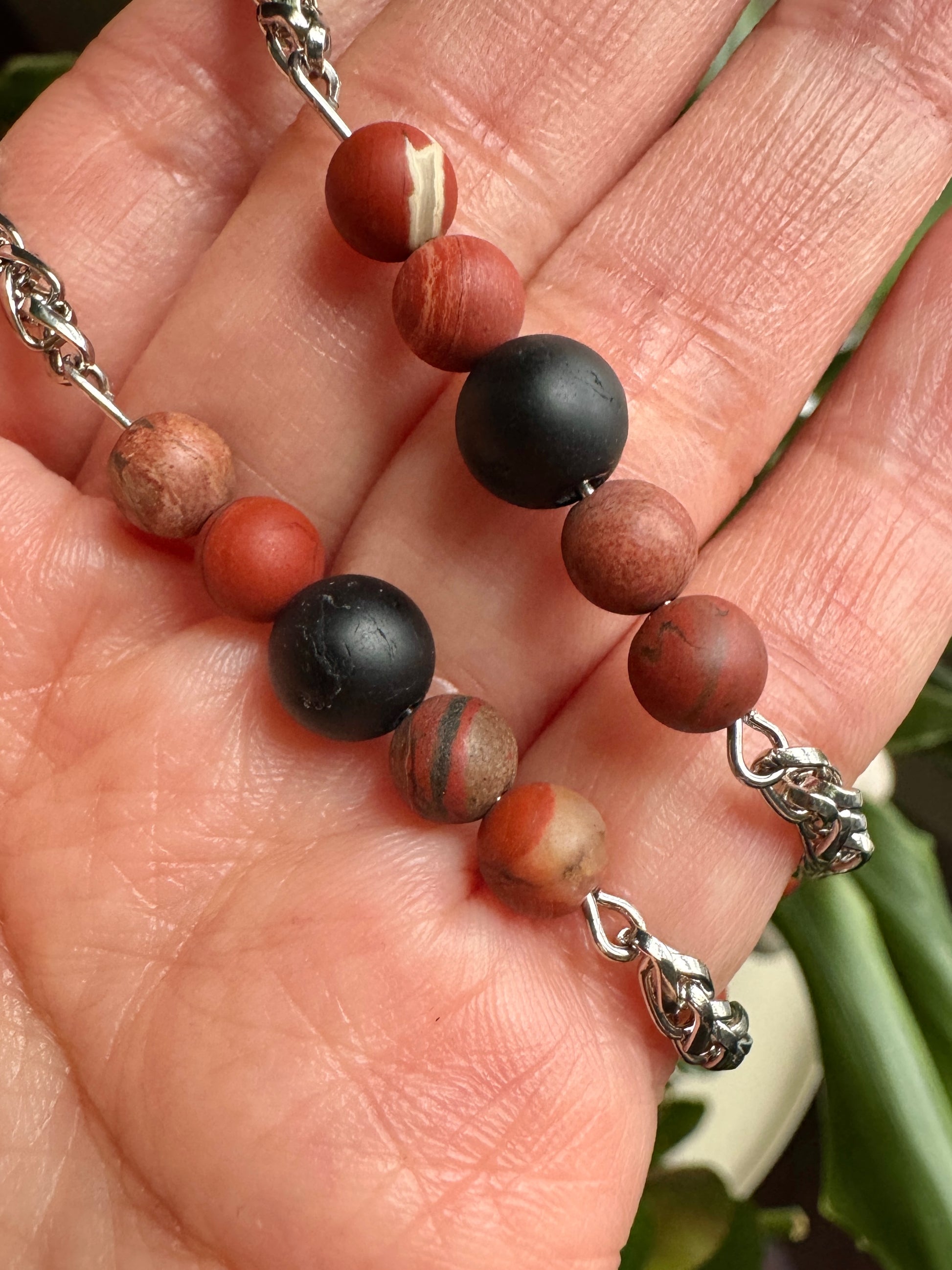 Hand holding a beaded bracelet with brown, black, and red beads on a blurred natural background