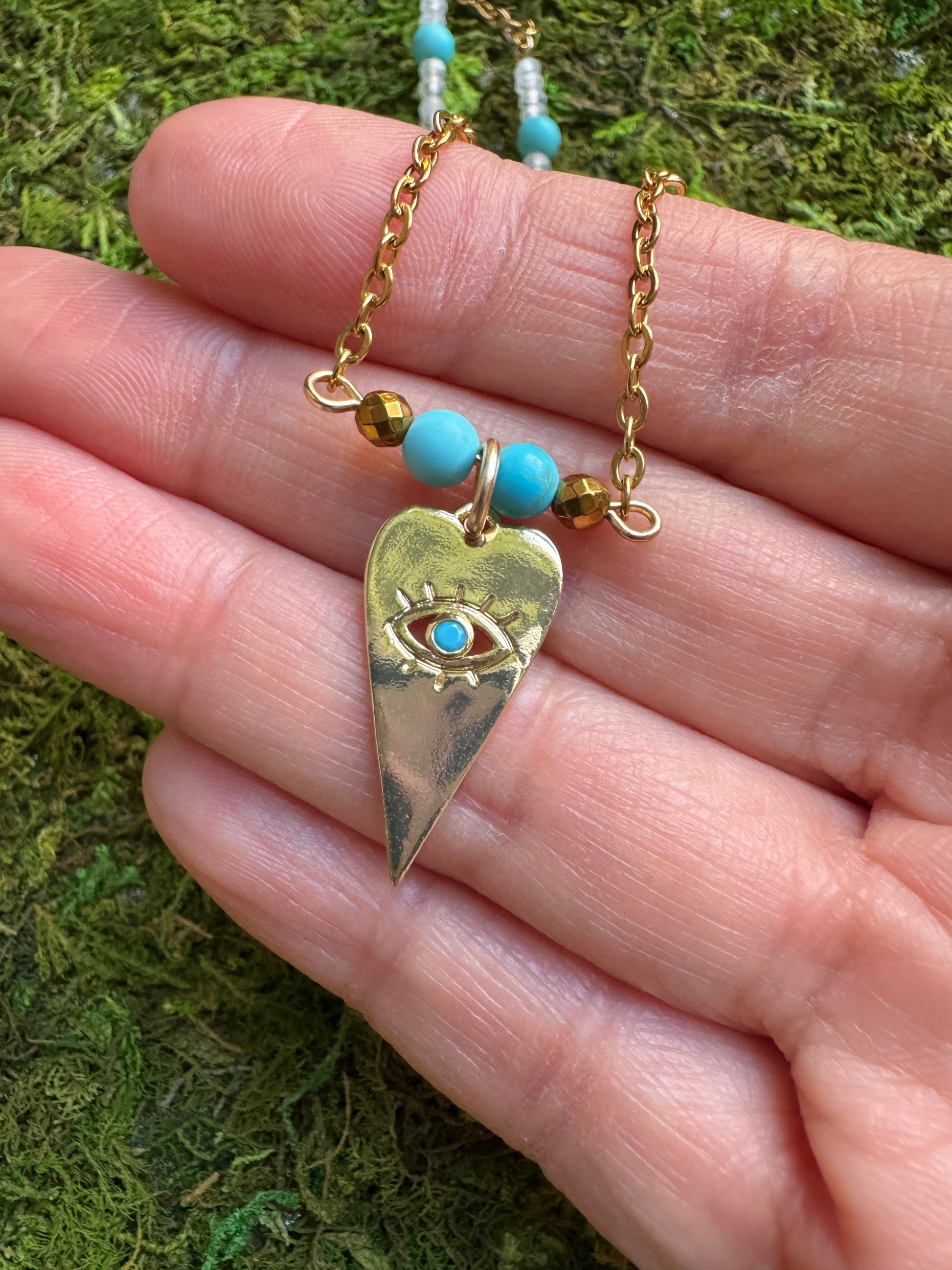 Gold necklace with a heart-shaped pendant featuring an eye design, held against a mossy background.