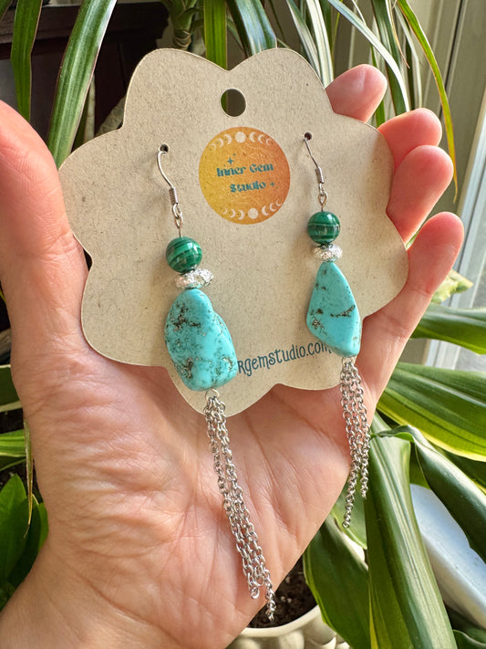 A hand holds a flower-shaped display card featuring a pair of turquoise and green beaded dangle earrings with silver chain drops, in front of green houseplant leaves. The display card has a circular logo for 'Anker Gold Studio'.