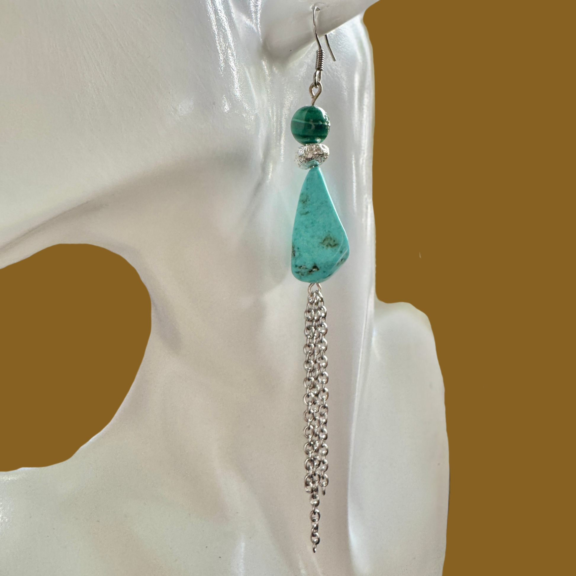 A single long dangle earring displayed on a white mannequin head against a warm brown background. The earring features a large turquoise-colored teardrop stone with dark matrix, a smaller dark green bead, a silver spacer ring, and a dangling silver chain.