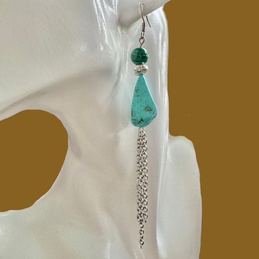 A single long dangle earring displayed on a white mannequin head against a warm brown background. The earring features a large turquoise-colored teardrop stone with dark matrix, a smaller dark green bead, a silver spacer ring, and a dangling silver chain.