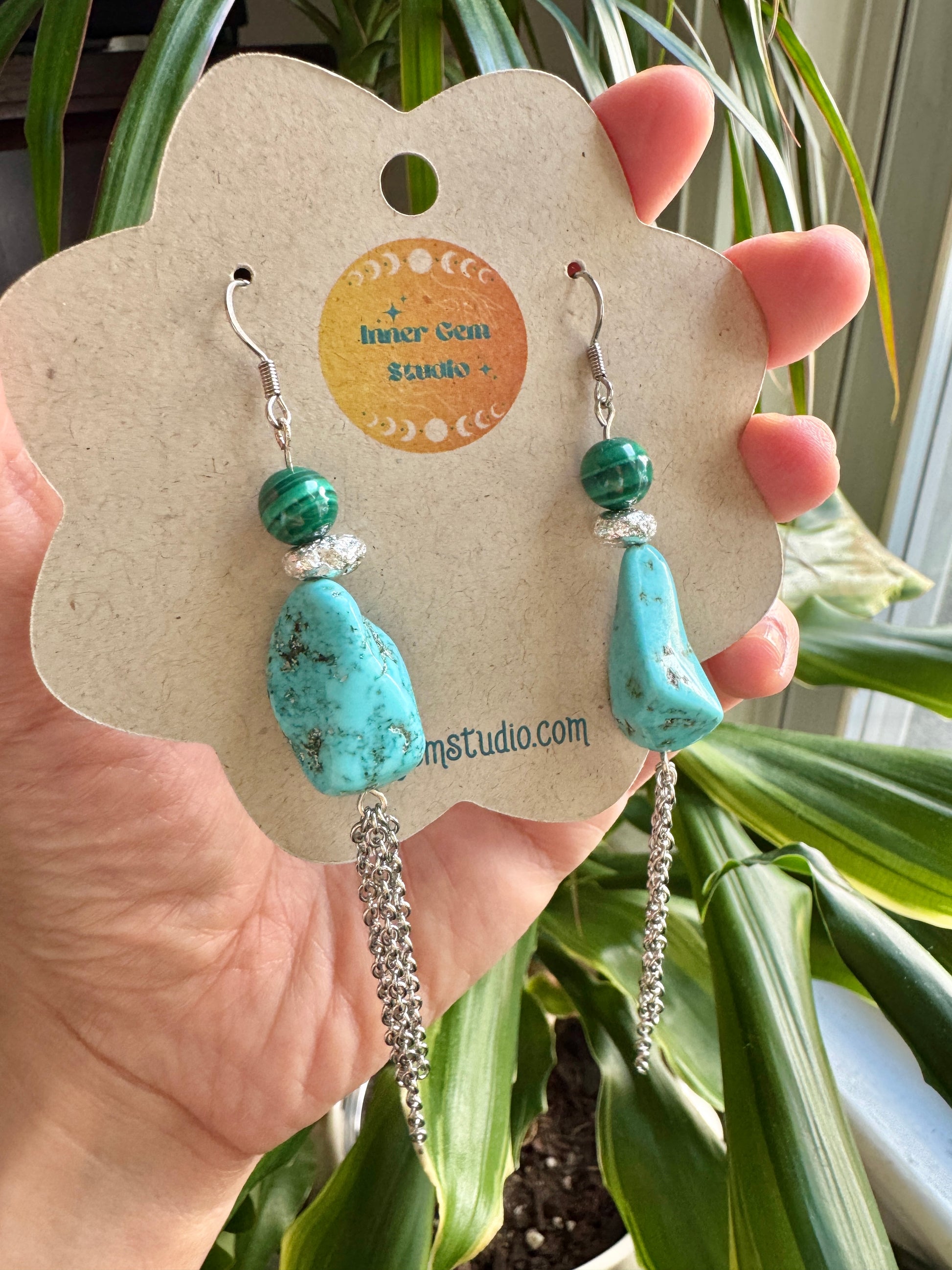 A hand holds a flower-shaped display card featuring a pair of turquoise and green beaded dangle earrings with silver chain drops, in front of green houseplant leaves.