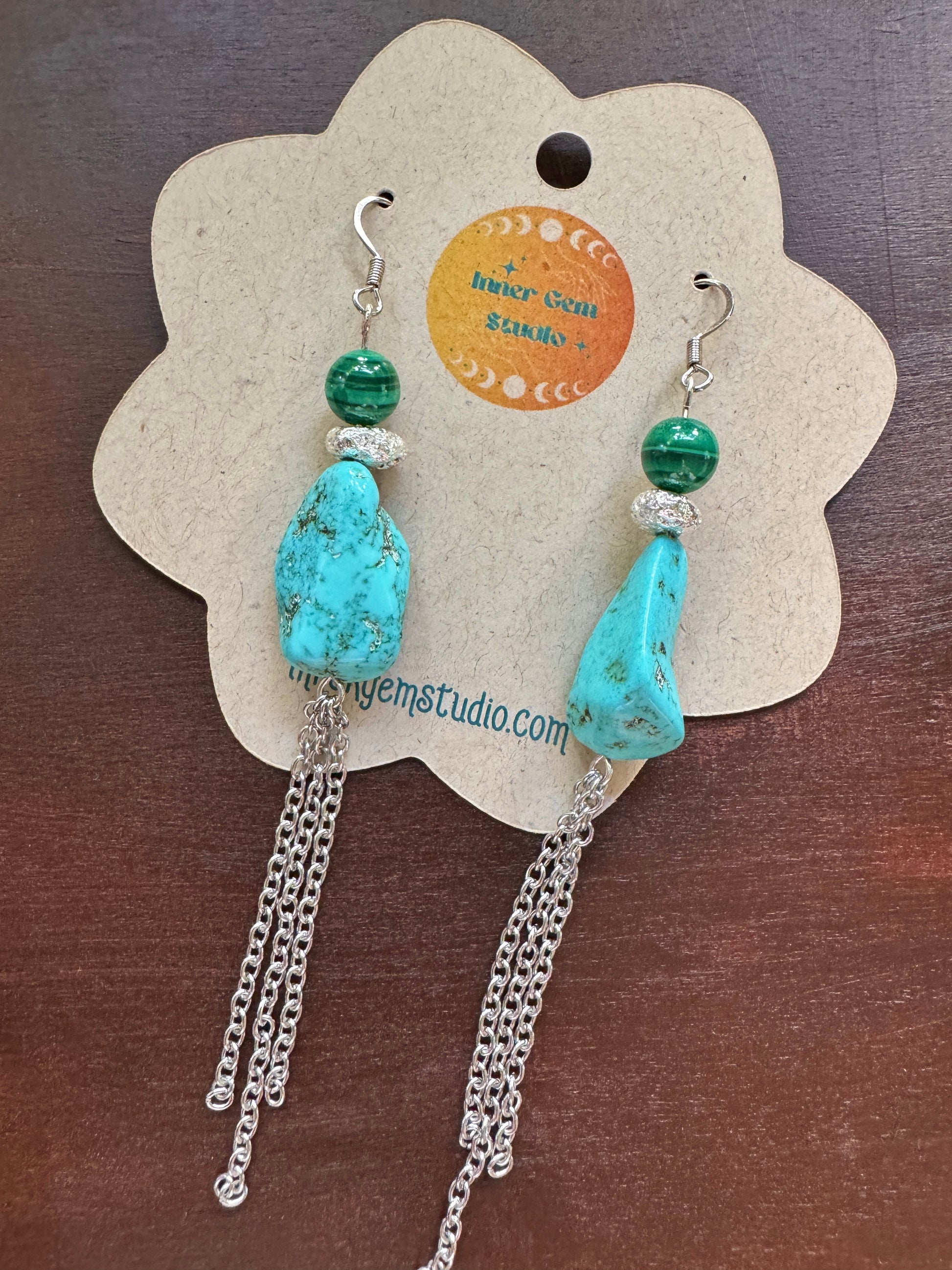 A pair of bohemian dangle earrings with turquoise and green beads, displayed on a flower-shaped kraft paper card. The earrings feature a large turquoise stone, a smaller green bead, a silver crystal accent, and dangling silver chains.